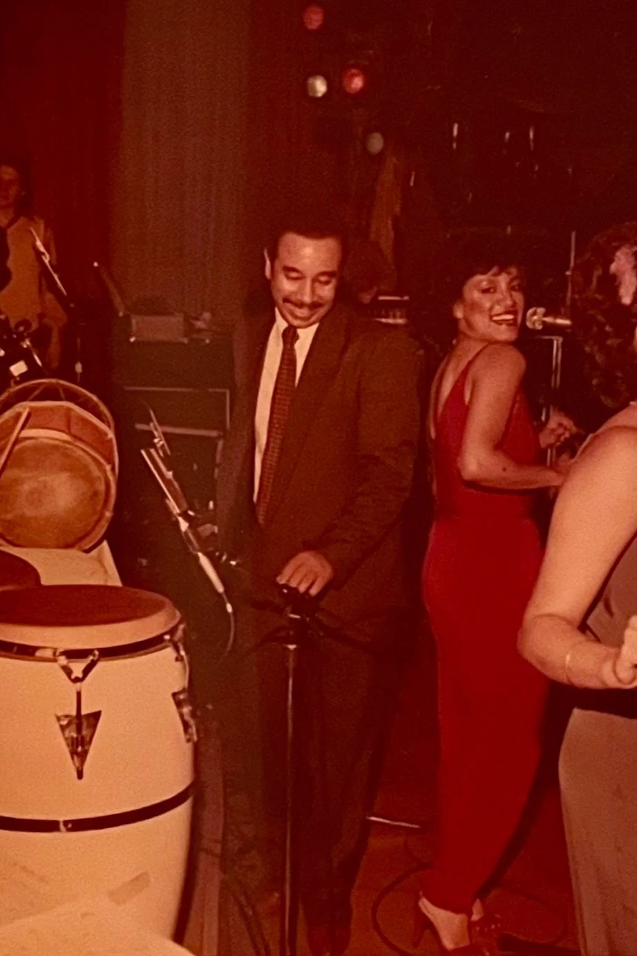 Vintage red-tinted photo of a man in a suit dancing beside a smiling woman in a red dress, with conga drums in the foreground.