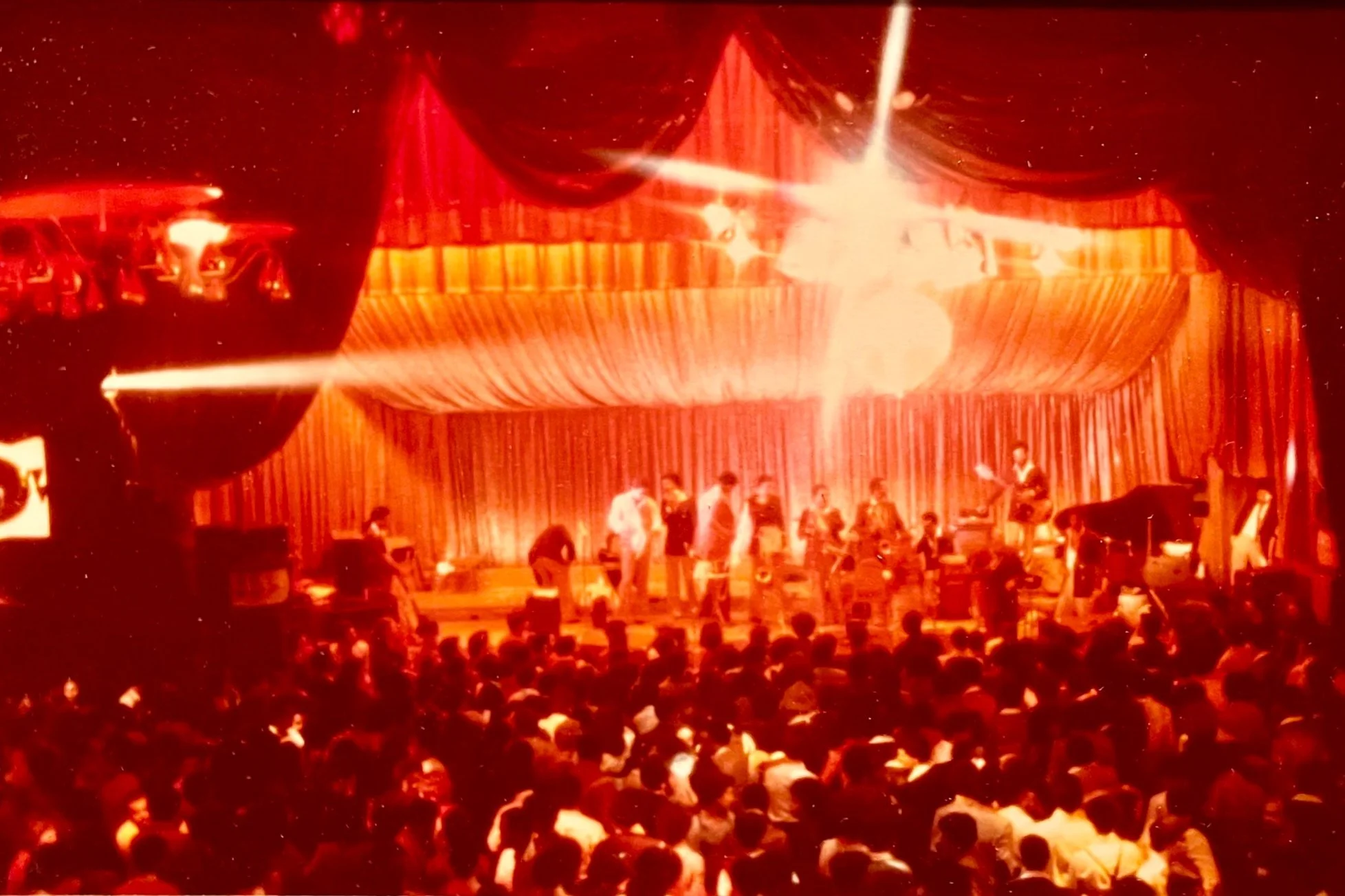 Vintage red-tinted photograph of a live band performing on a decorated stage before a large audience