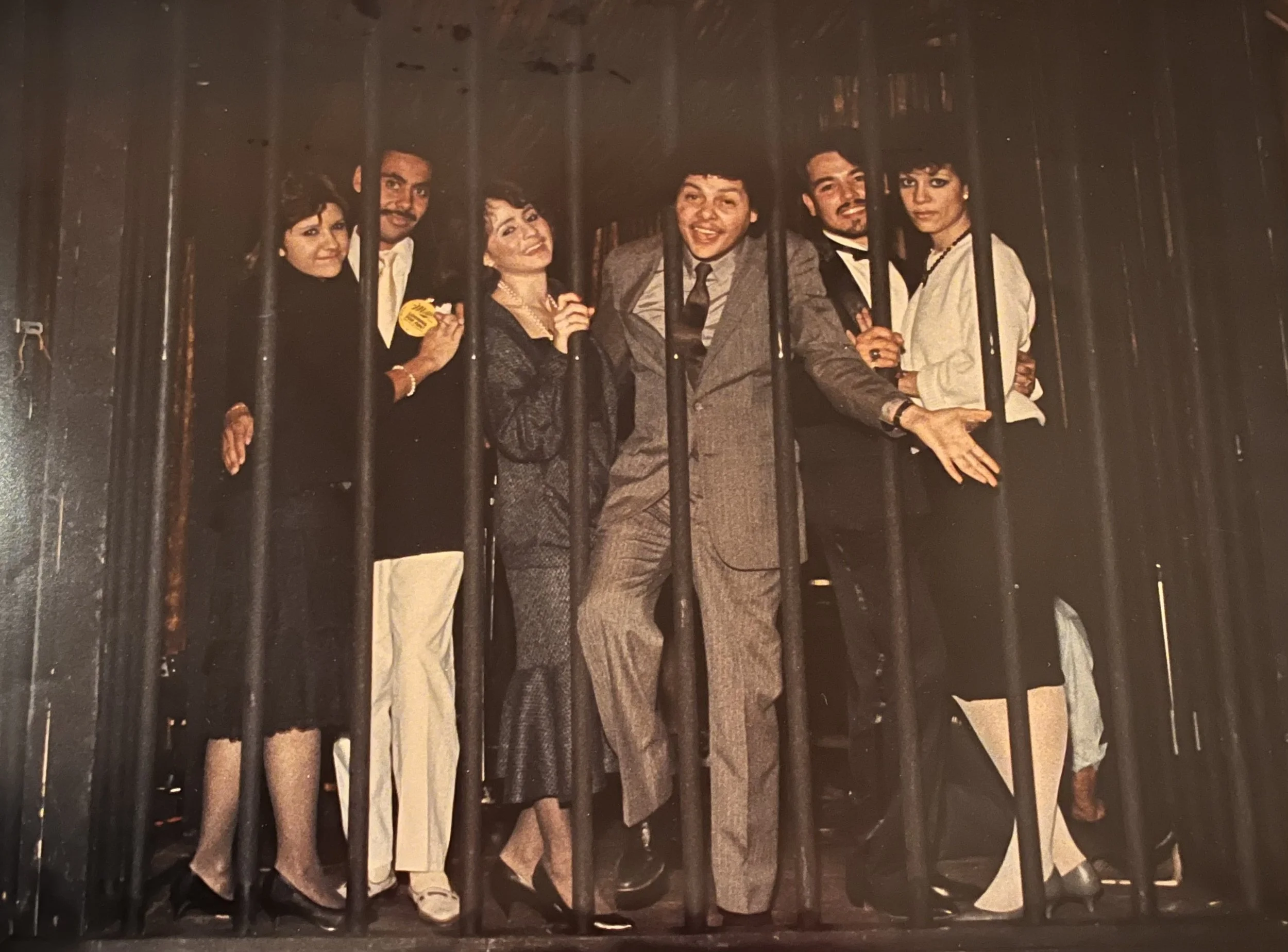 Vintage group portrait of six people standing behind vertical metal bars, dressed in formal and theatrical clothing.