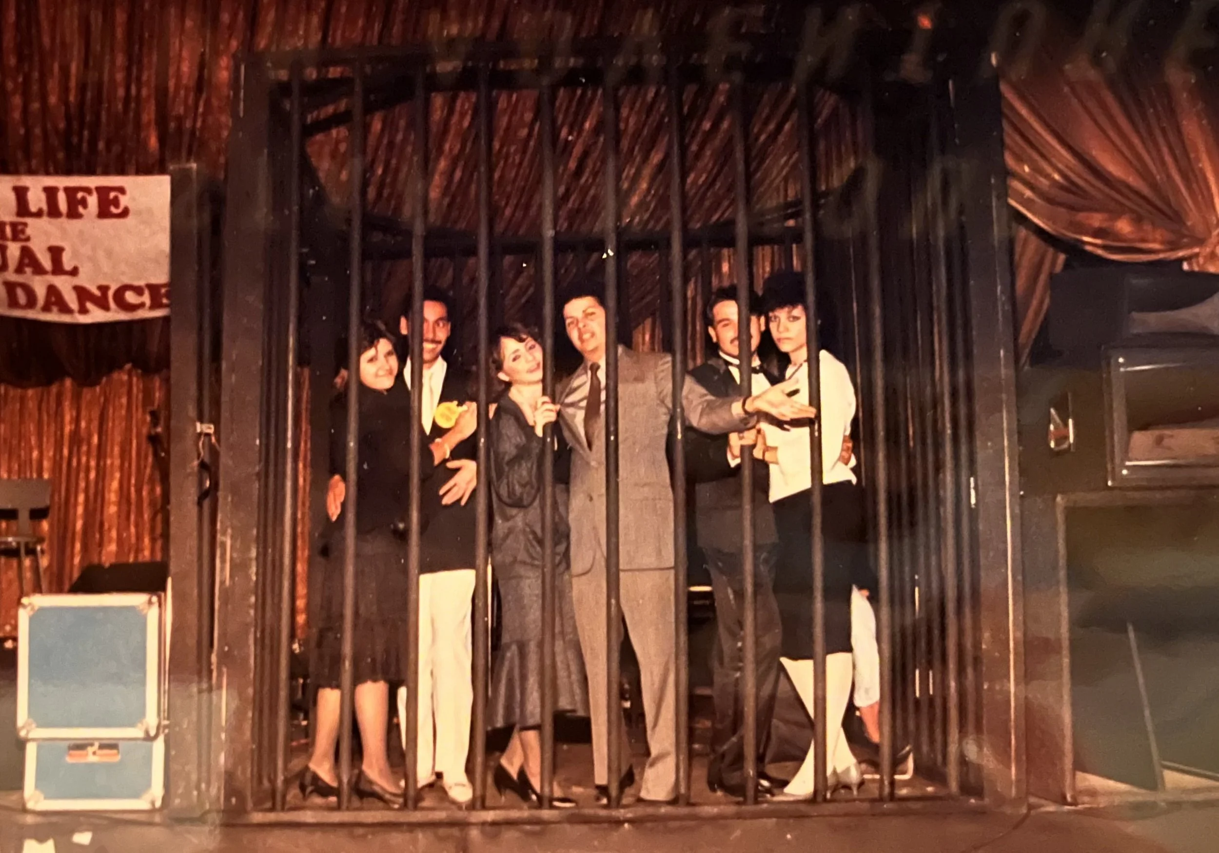 Theater performers pose behind prison bars on a dimly lit stage, with a vintage sign reading “Life … Dance.”