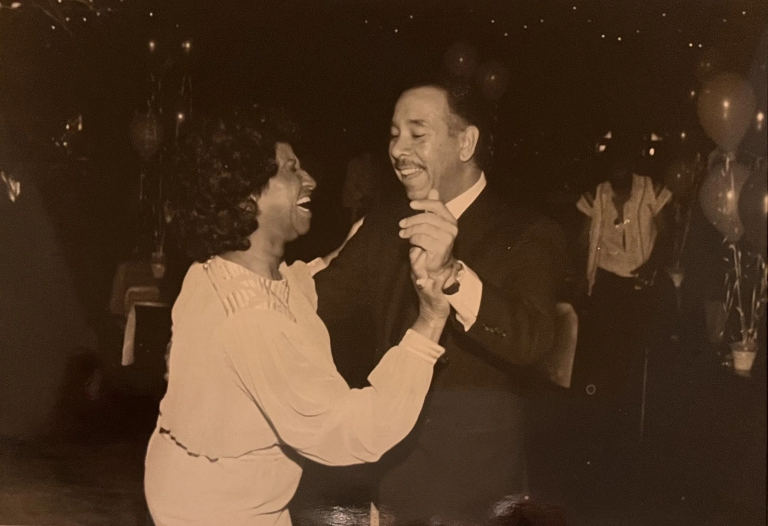 Sepia-toned photo of a smiling couple dancing at a formal evening event as guests applaud.