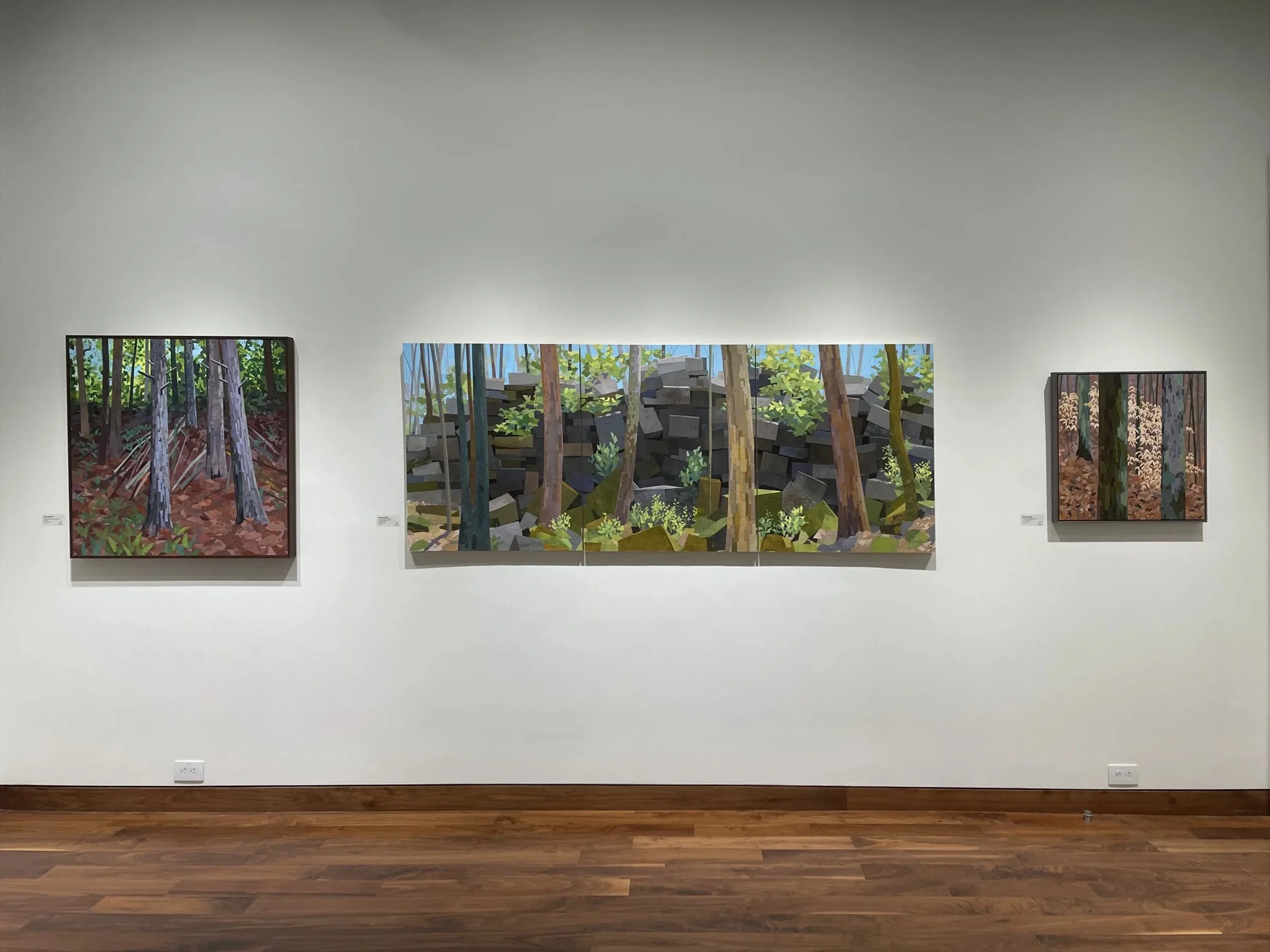 Three landscape paintings in a gallery, featuring trees, rocks, and forest scenes, displayed on a white wall with wooden floor.