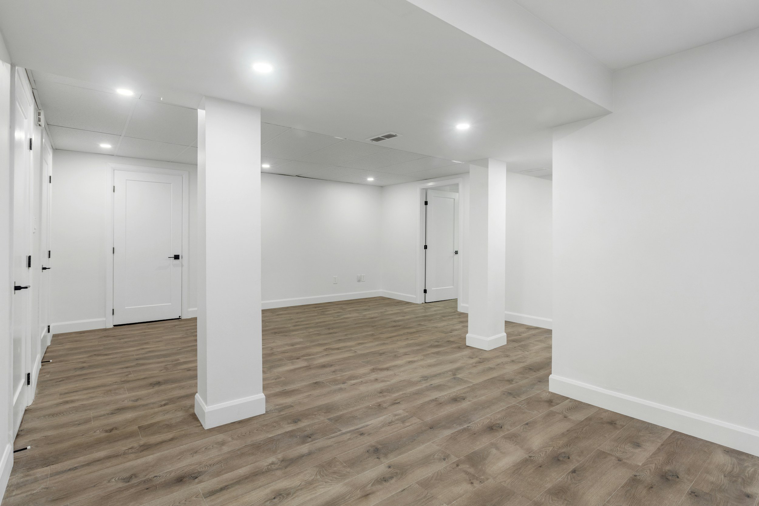 Empty basement room with white walls, white ceiling, wood laminate flooring, and multiple white doors, illuminated by recessed ceiling lights.