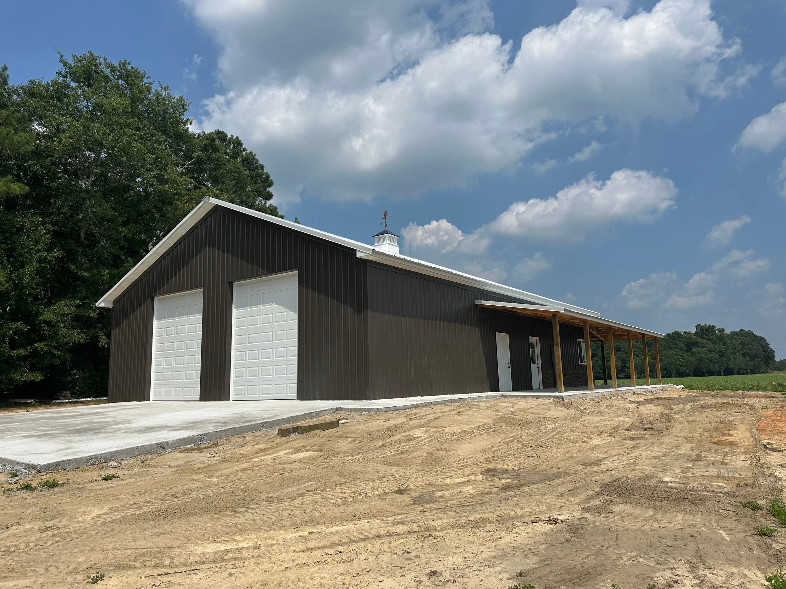 A black metal building with two white garage doors and a small porch area with wooden posts, situated on a dirt lot with a concrete pad, under a partly cloudy sky.