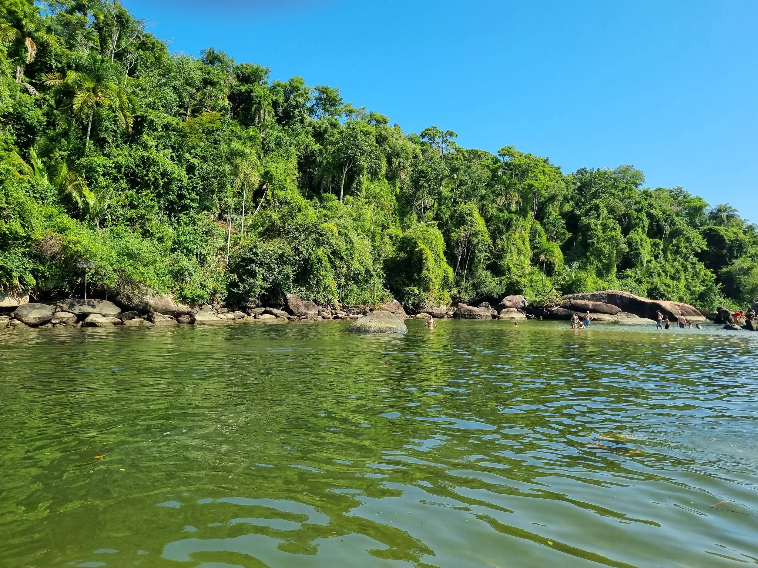 Rio de água clara com pessoas nadando e uma encosta coberta por árvores verdes sob céu azul