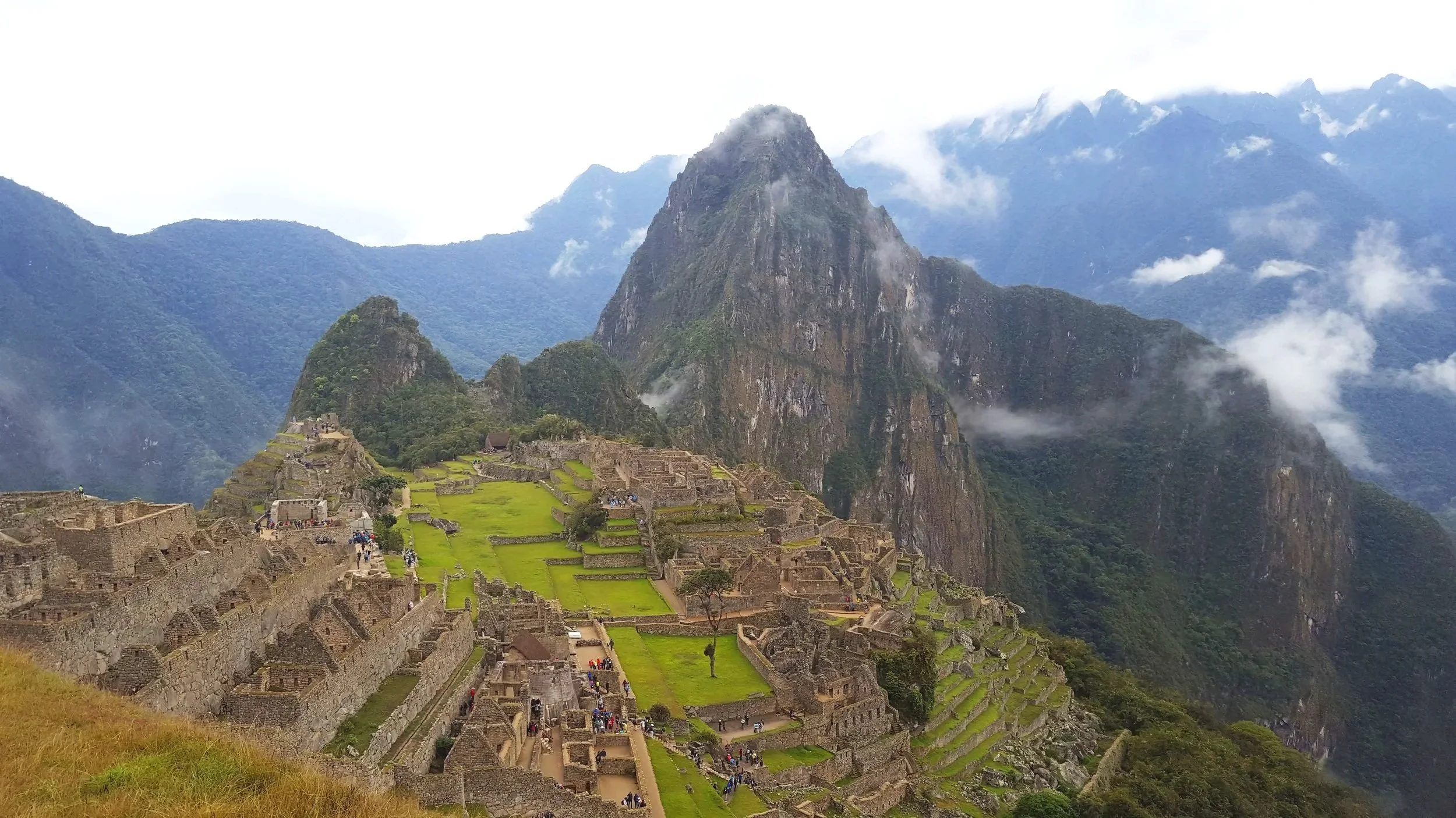 Paisagem de Machu Picchu com ruínas antigas, vegetação verdejada e montanhas ao fundo, incluindo o Aguja Peak, cercado por nuvens.
