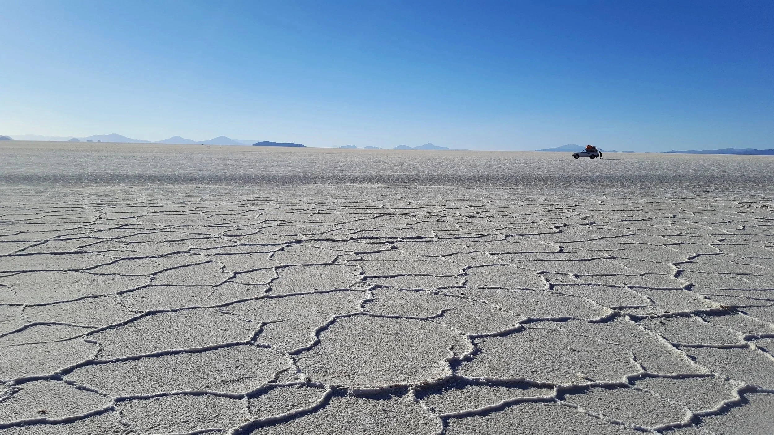 Paisagem de um deserto com solo rachado, céu azul claro e uma caminhonete com equipamento no topo ao longe, em um ambiente seco e árido.