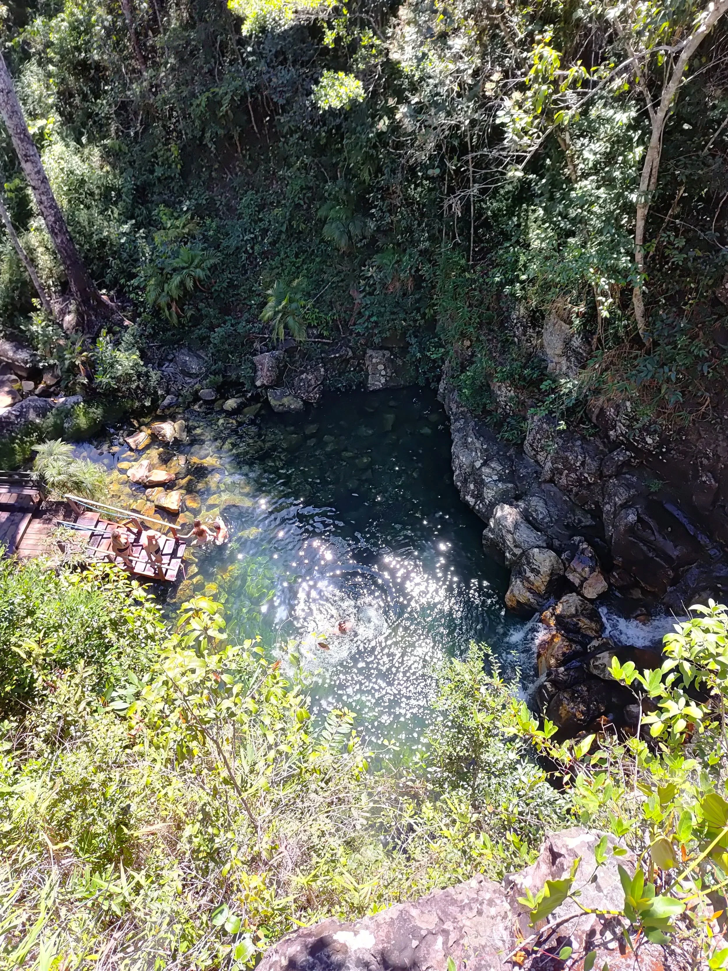 Foto de uma nascente de água natural cercada por vegetação densa e rochas, com algumas pessoas ao lado, na parte inferior esquerda.