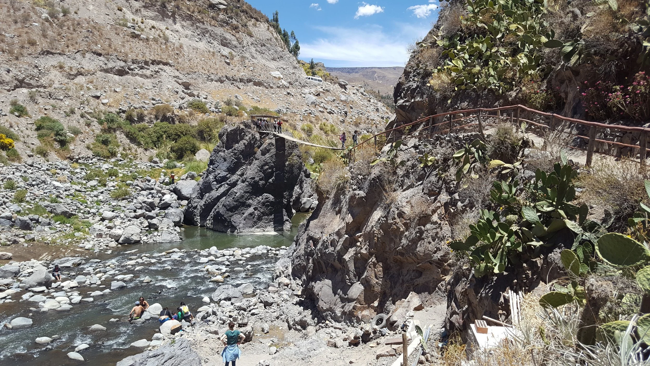 Cachoeira de rio cercada por rochas e vegetação, com uma ponte suspensa de madeira sobre o rio, e pessoas aproveitando a cachoeira em um dia de clima ensolarado