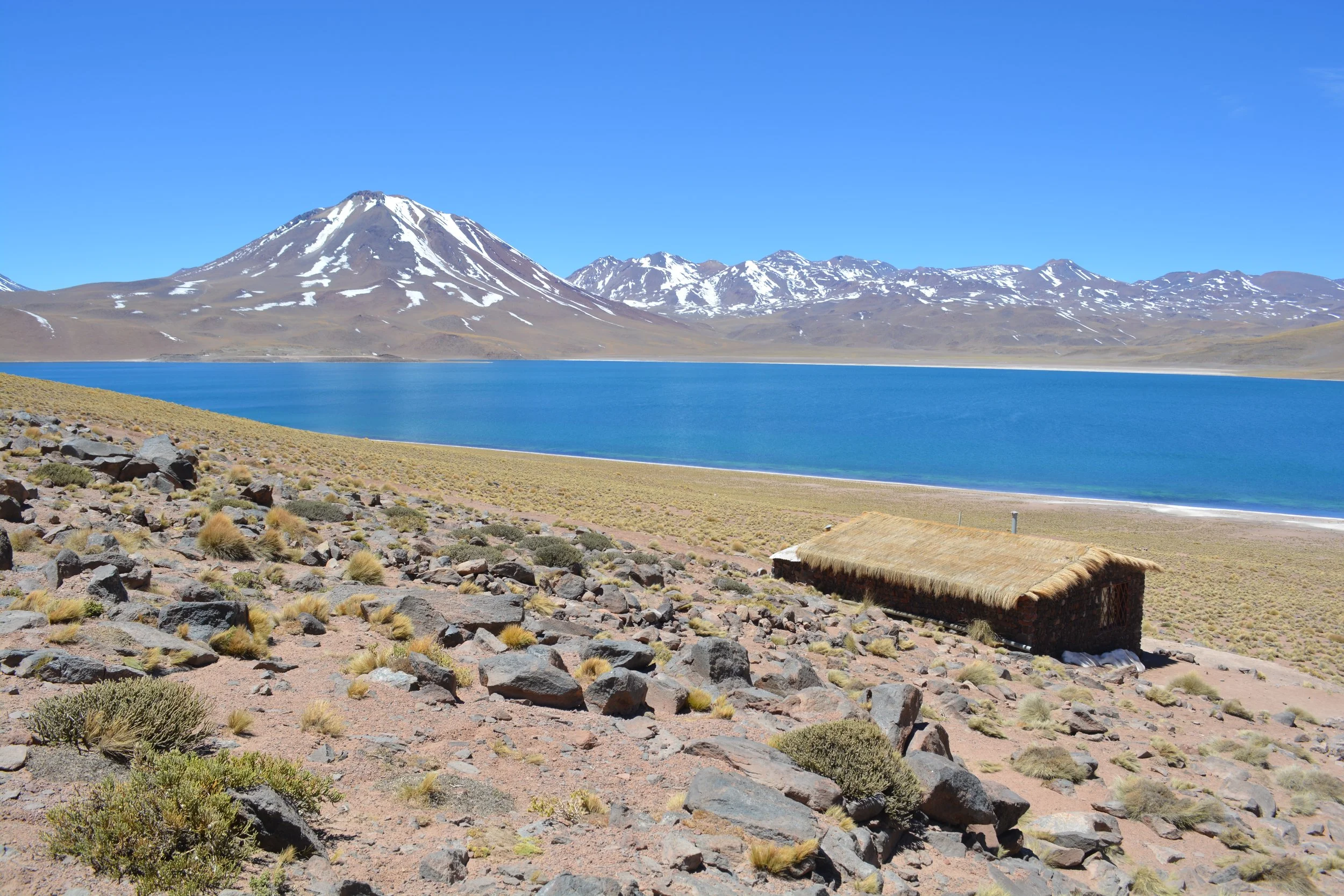 Montanhas nevadas ao fundo, lago azul em primeiro plano, terra árida com pedras e mato, uma cabana de palha no canto inferior direito.