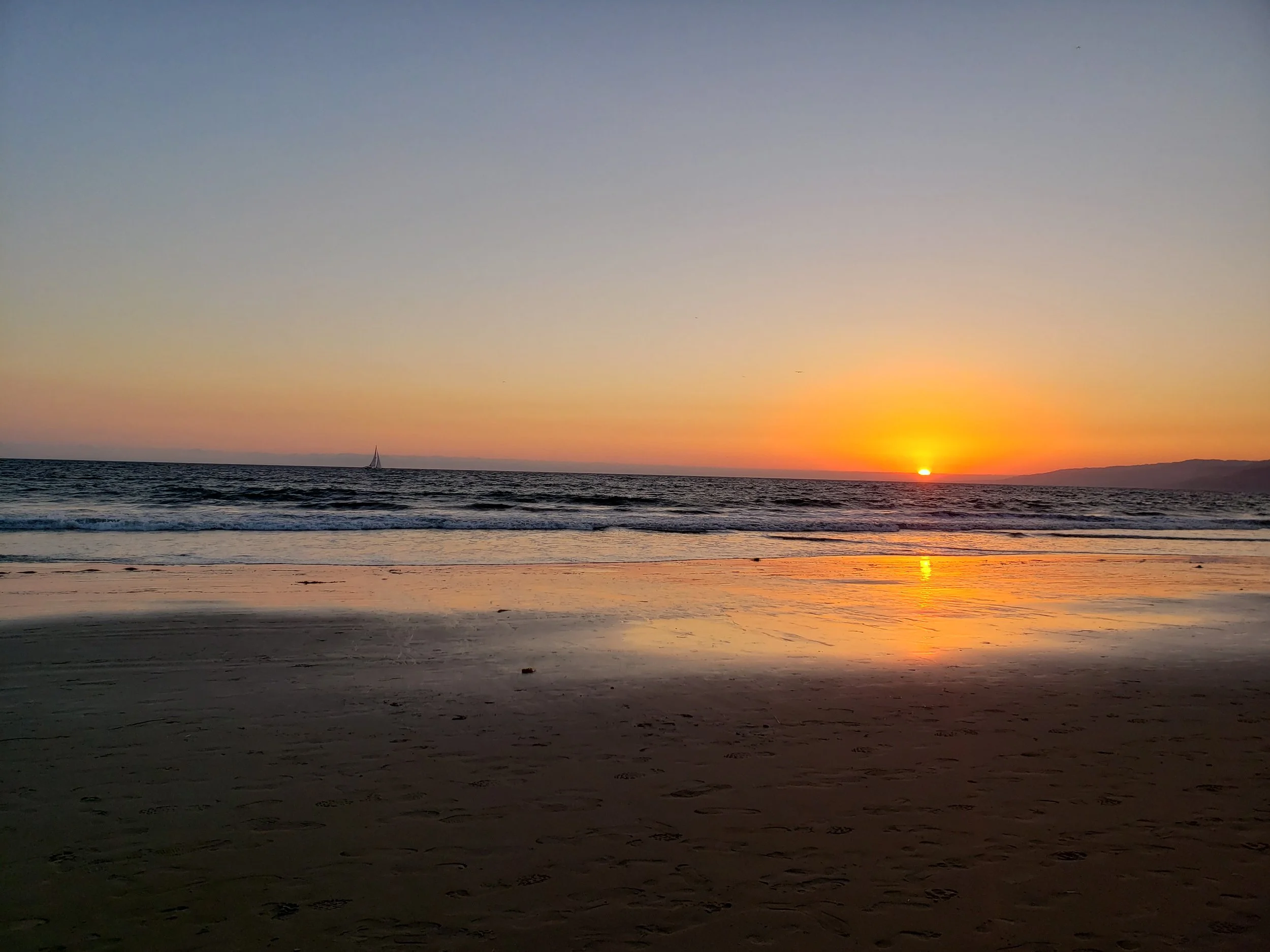 Pôr do sol na praia com céu em tons de laranja, roxo e azul, mar calmo e uma vela ao longe.