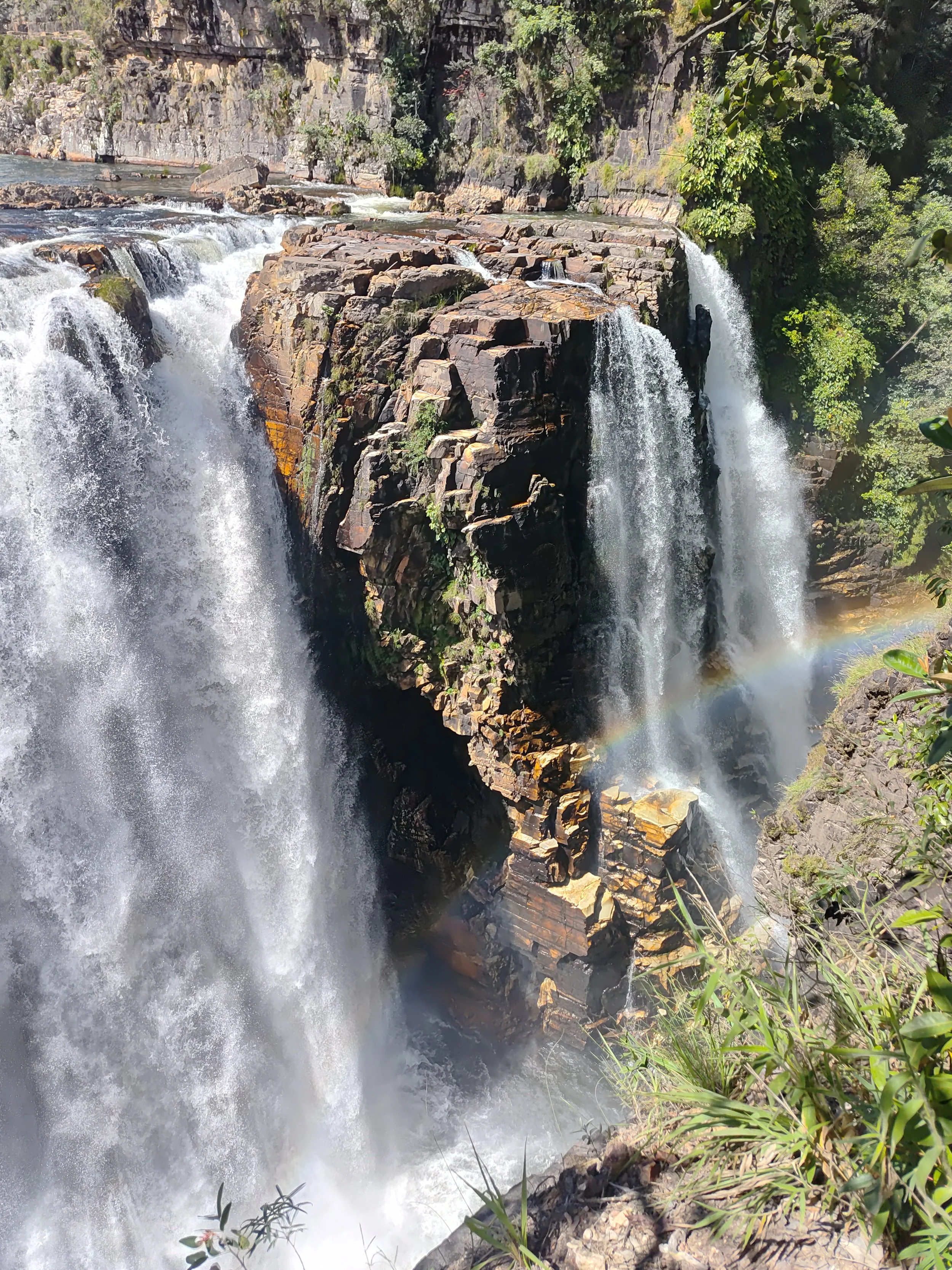 Cachoeira com água caindo sobre rochas, cercada por vegetação verde, com arco-íris visível na água spray.