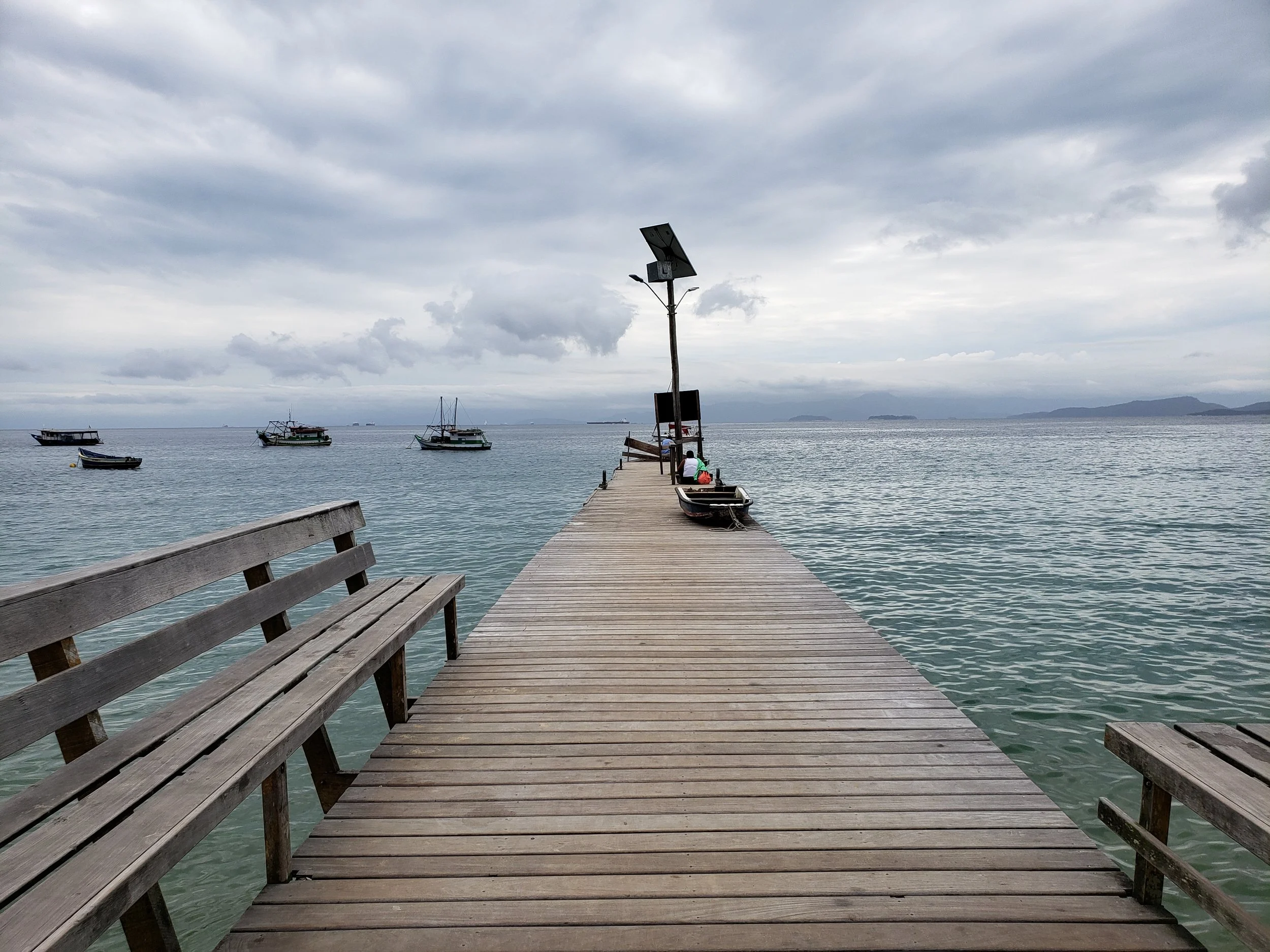 Píer de madeira com bancos de um lado, estende-se sobre águas calmas, com barcos ancorados ao redor e céu nublado ao fundo.