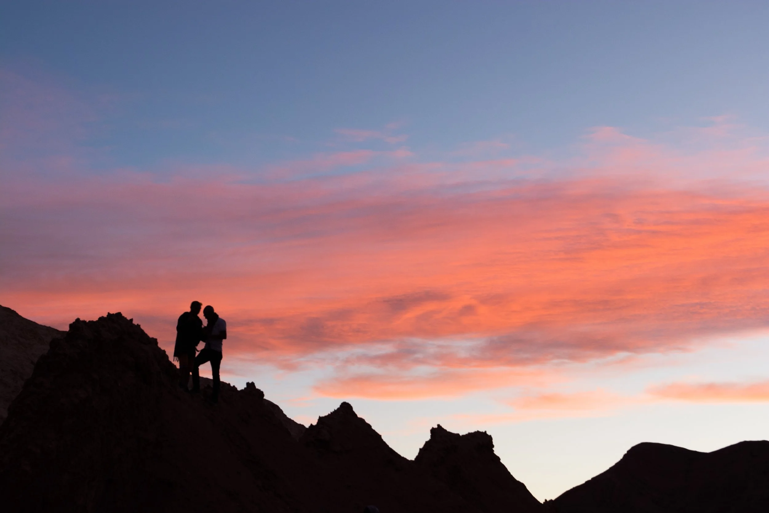 Homens em pé sobre uma formação rochosa olhando para o horizonte, durante o pôr do sol com céu colorido em tons de rosa, azul e laranja.