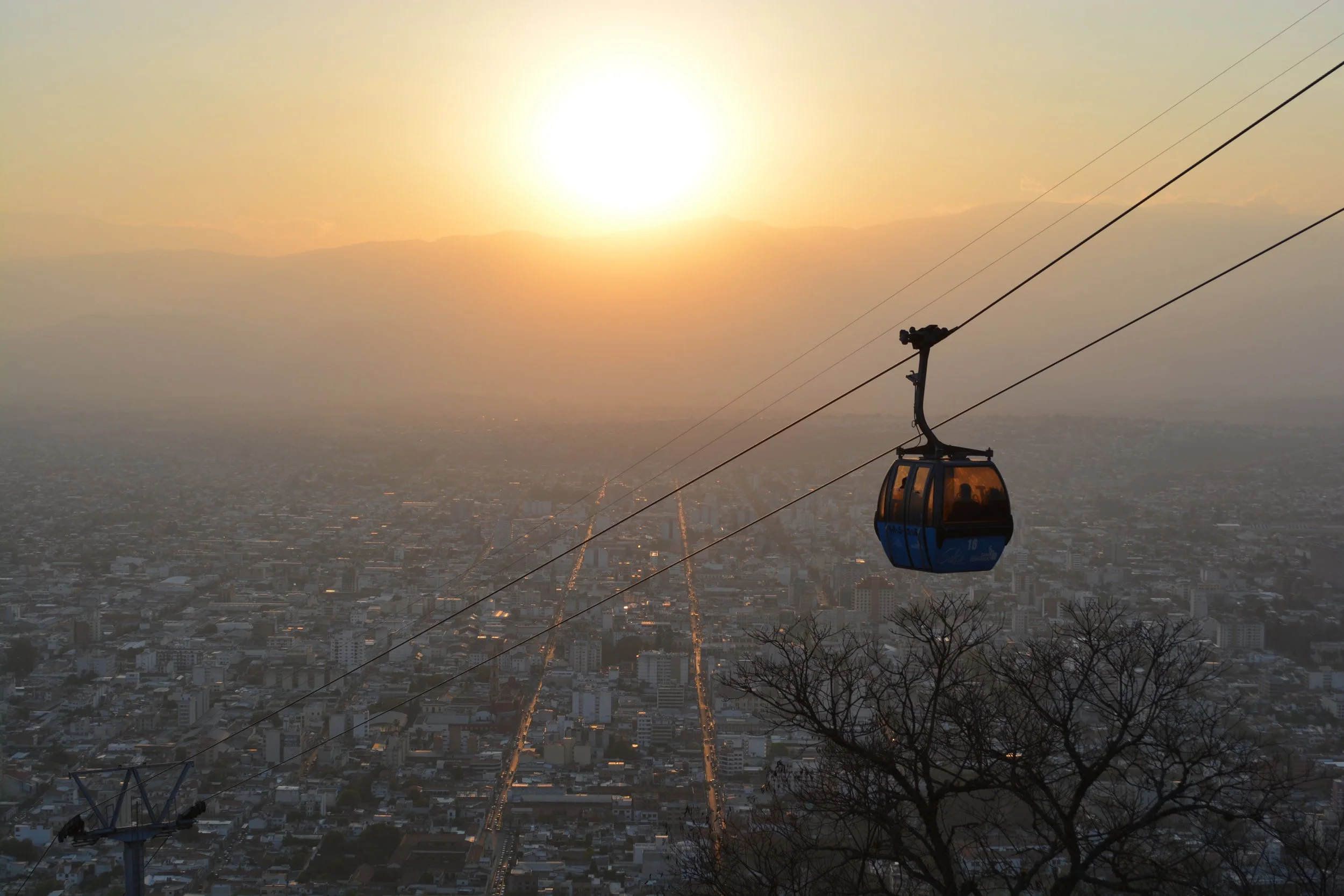 Câmbio de teleférico sobre uma cidade ao entardecer, com o sol se pondo no horizonte, e montanhas ao fundo.
