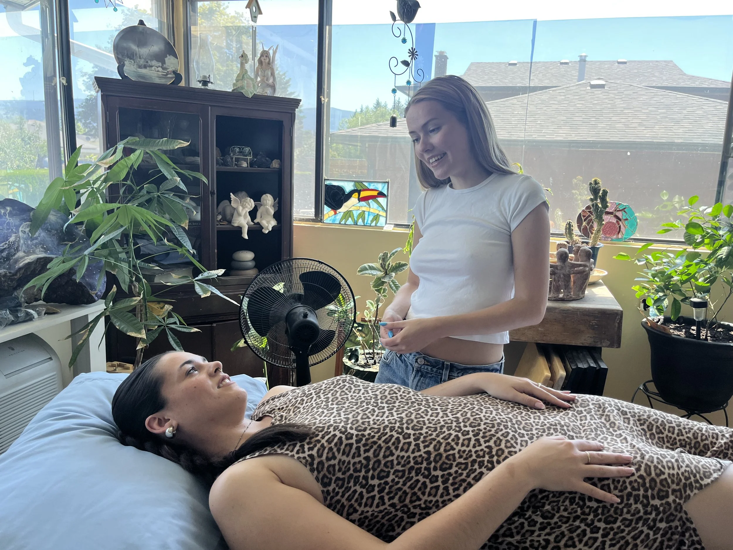 Two young women are in a bright, sunlit room filled with plants and decorative items. One woman is lying on a bed with a leopard print blanket, smiling and looking at the other woman who is standing and smiling at her. The room has a mix of vintage and eclectic decor, with plants, art, and figurines on shelves.