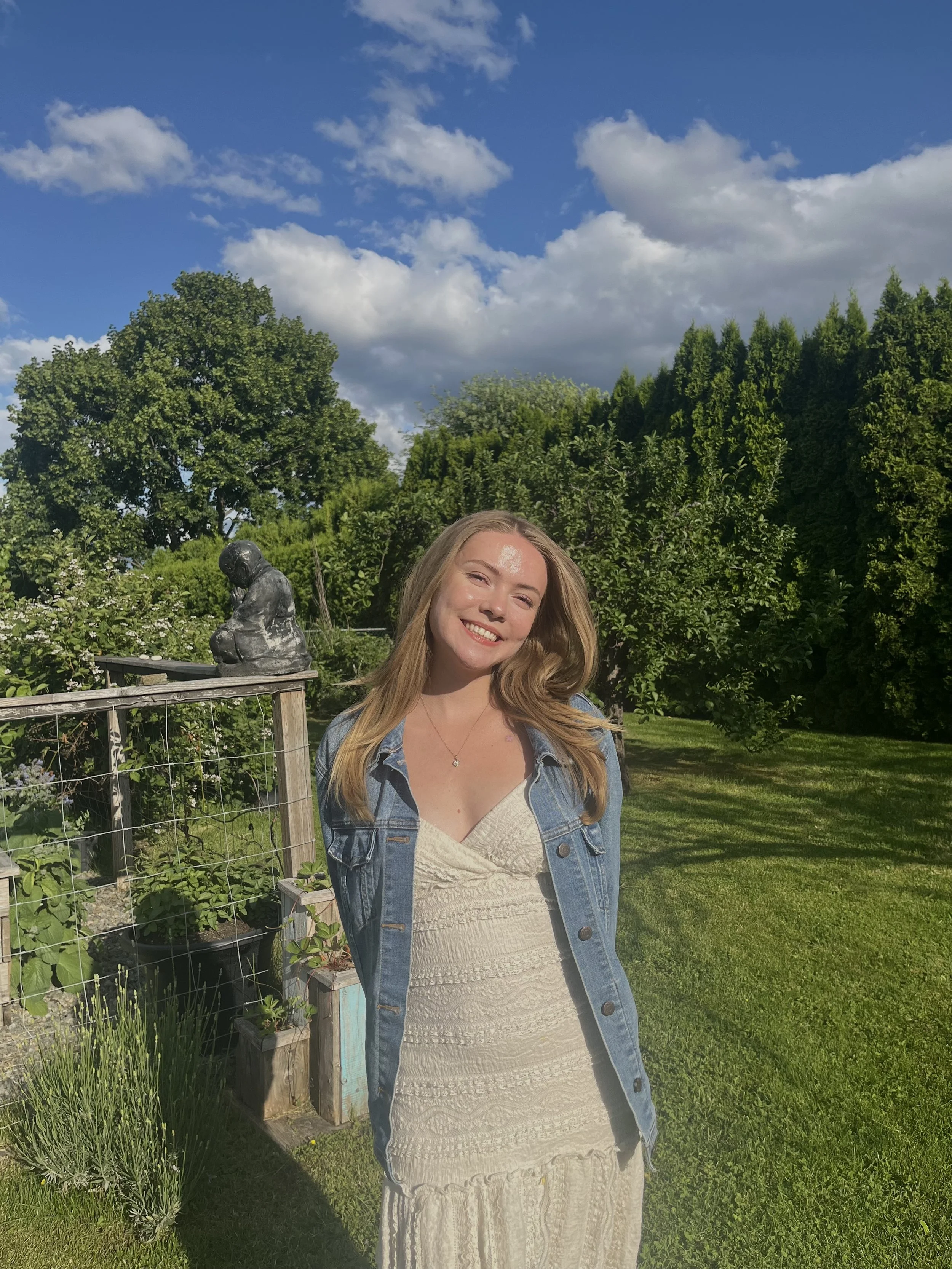 A young woman with long blonde hair smiling outdoors in a garden with trees, bushes, a wooden fence, and clouds in the sky.