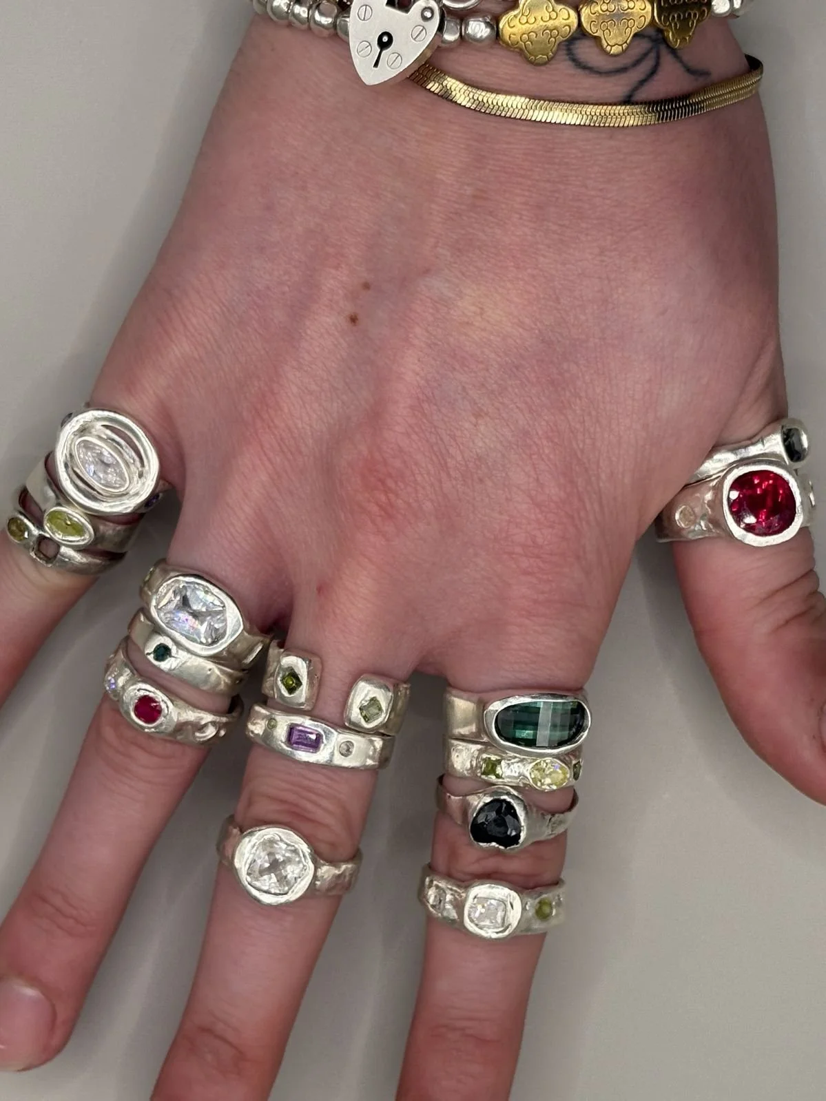 A hand wearing multiple silver rings with various colorful gemstones and a large dark green gemstone. The person is also wearing several necklaces, including a gold link chain, a heart-shaped gold pendant, a watch face, and a silver beaded bracelet.