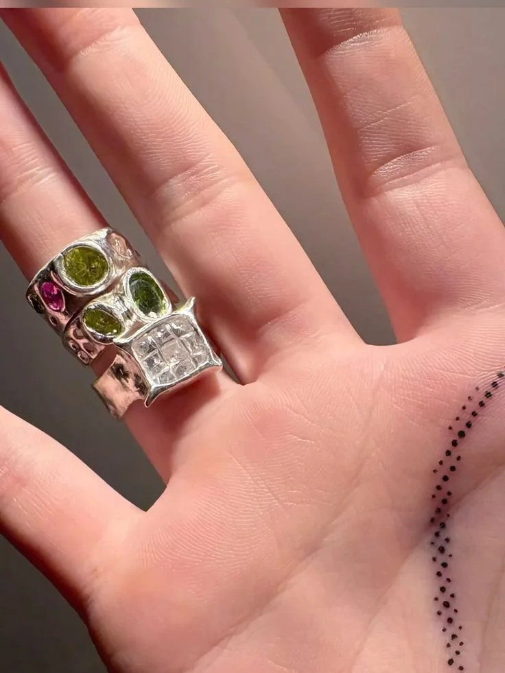 Close-up of a hand wearing a chunky silver ring with three embedded green gems, one small pink gem, and a square clear gemstone. The person has small black dotted tattoo on their finger and the background is plain gray.