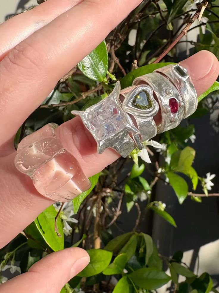 Multiple silver rings with gemstones, including one with a green heart-shaped gem, another with a red gemstone, and a clear ring with a square pattern, held on fingers against a leafy background.