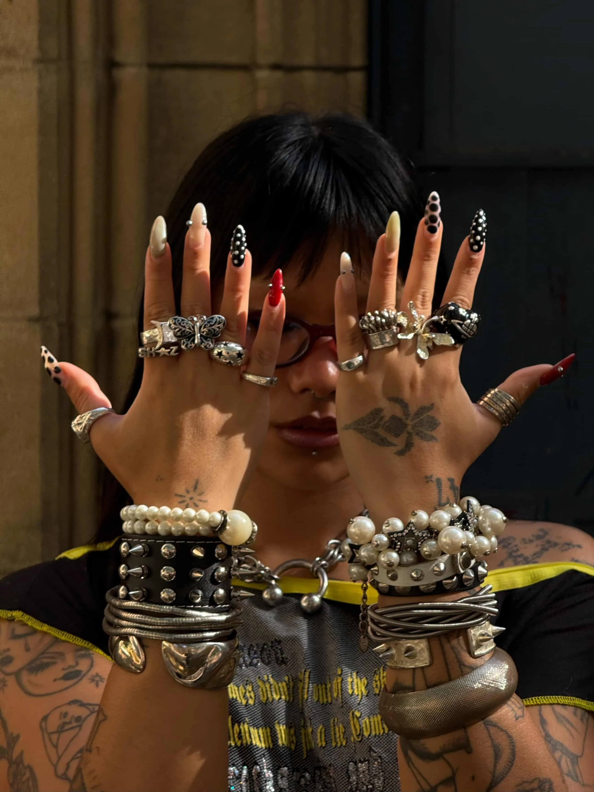 Person with black hair and glasses displaying hands covered in rings and bracelets, with tattoos on arms and hands, in a dimly lit outdoor setting.