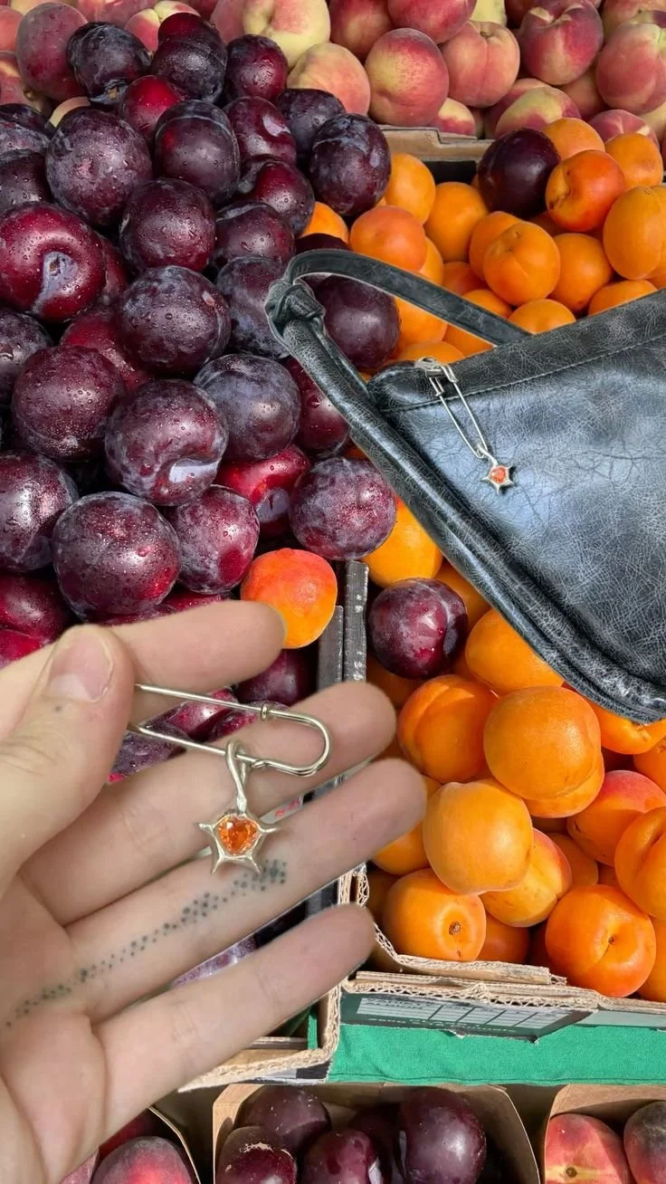 A display of fresh purple plums, orange apricots, and pink peaches at a grocery store produce section, with a black handbag and a person's hand holding jewelry.