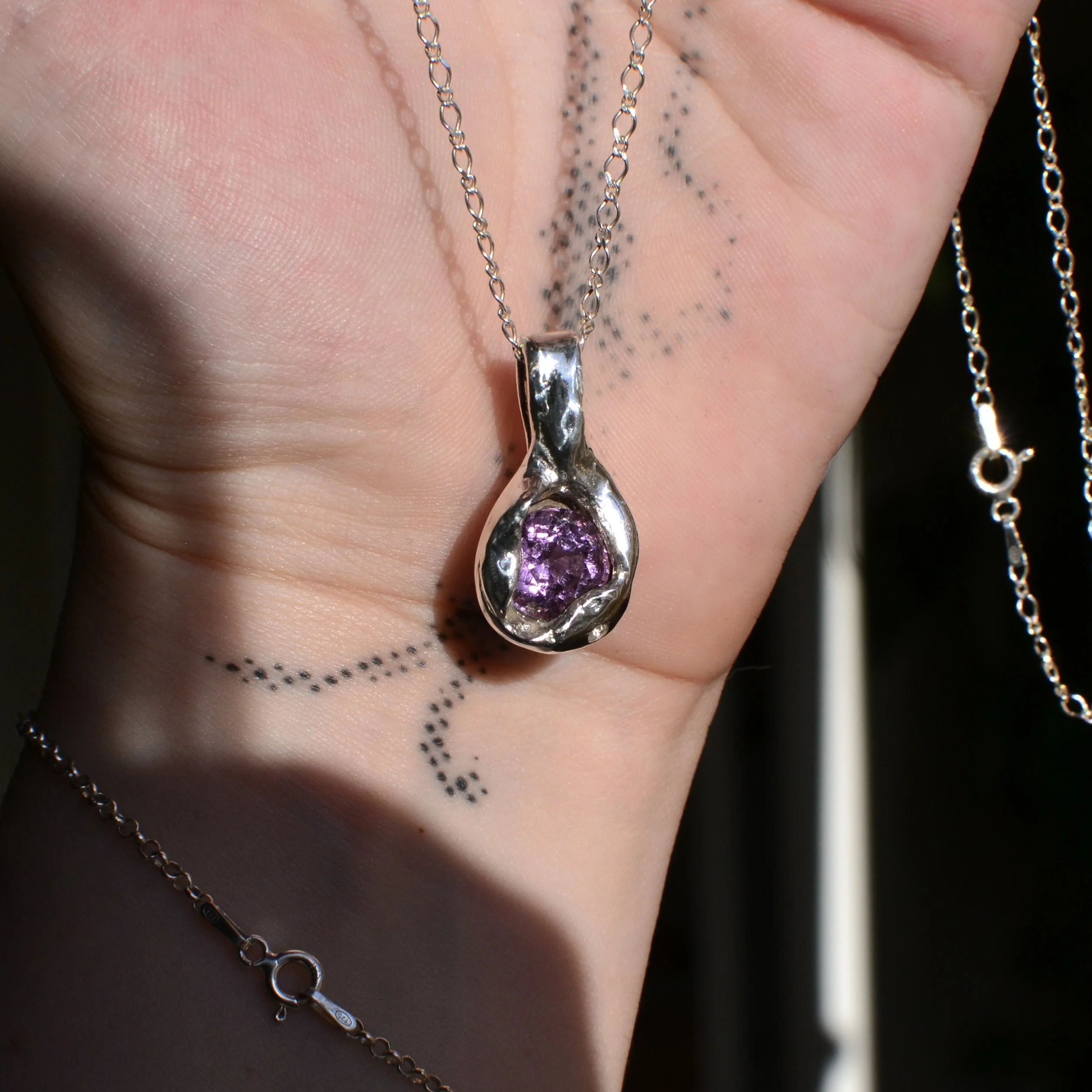 Silver necklace with a purple gemstone pendant held against a hand with dotted black tattoo lines.