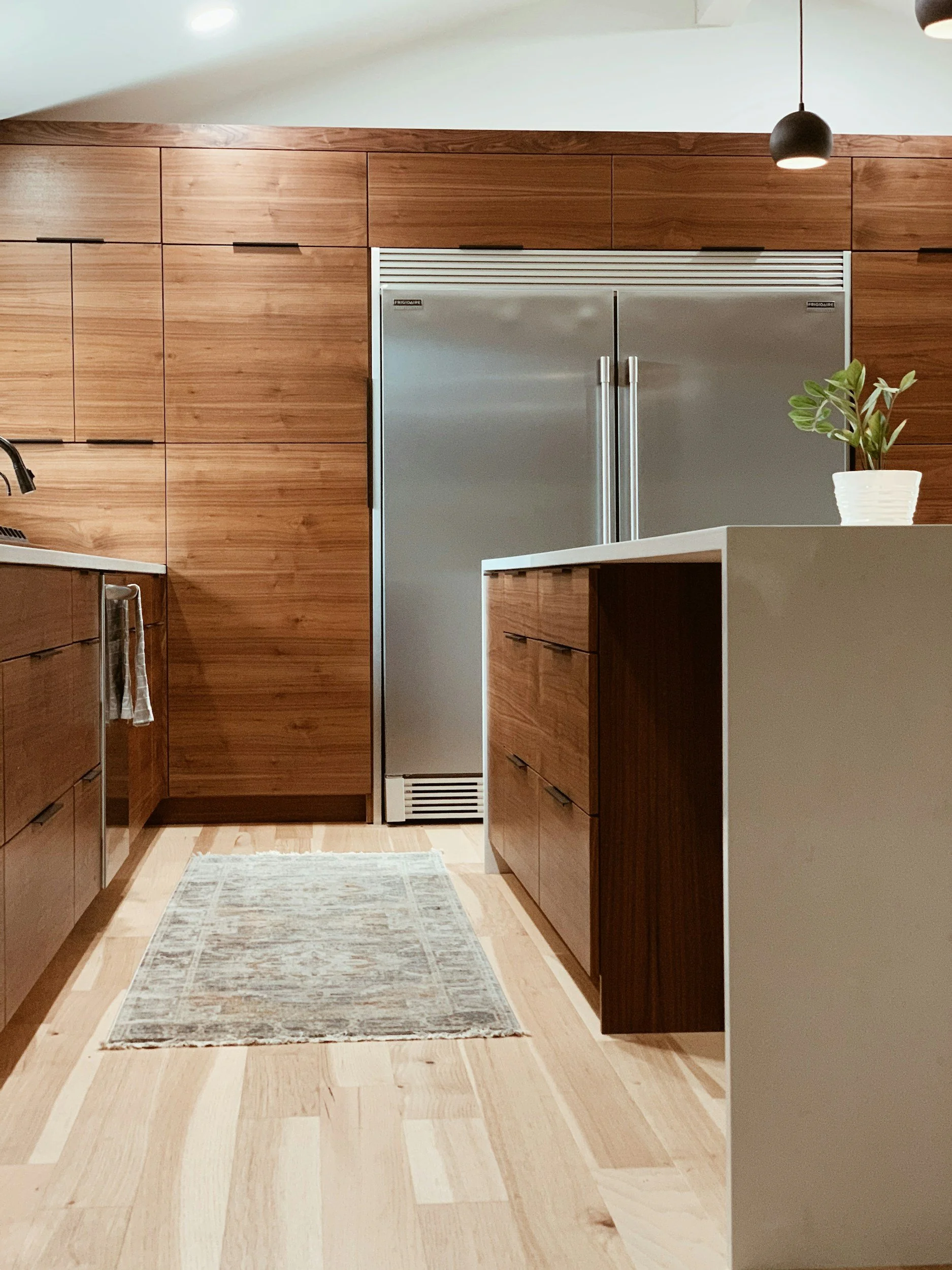 Modern kitchen with wooden cabinets, stainless steel refrigerator, a small rug on light wood flooring, and a white kitchen island with a potted plant on top.