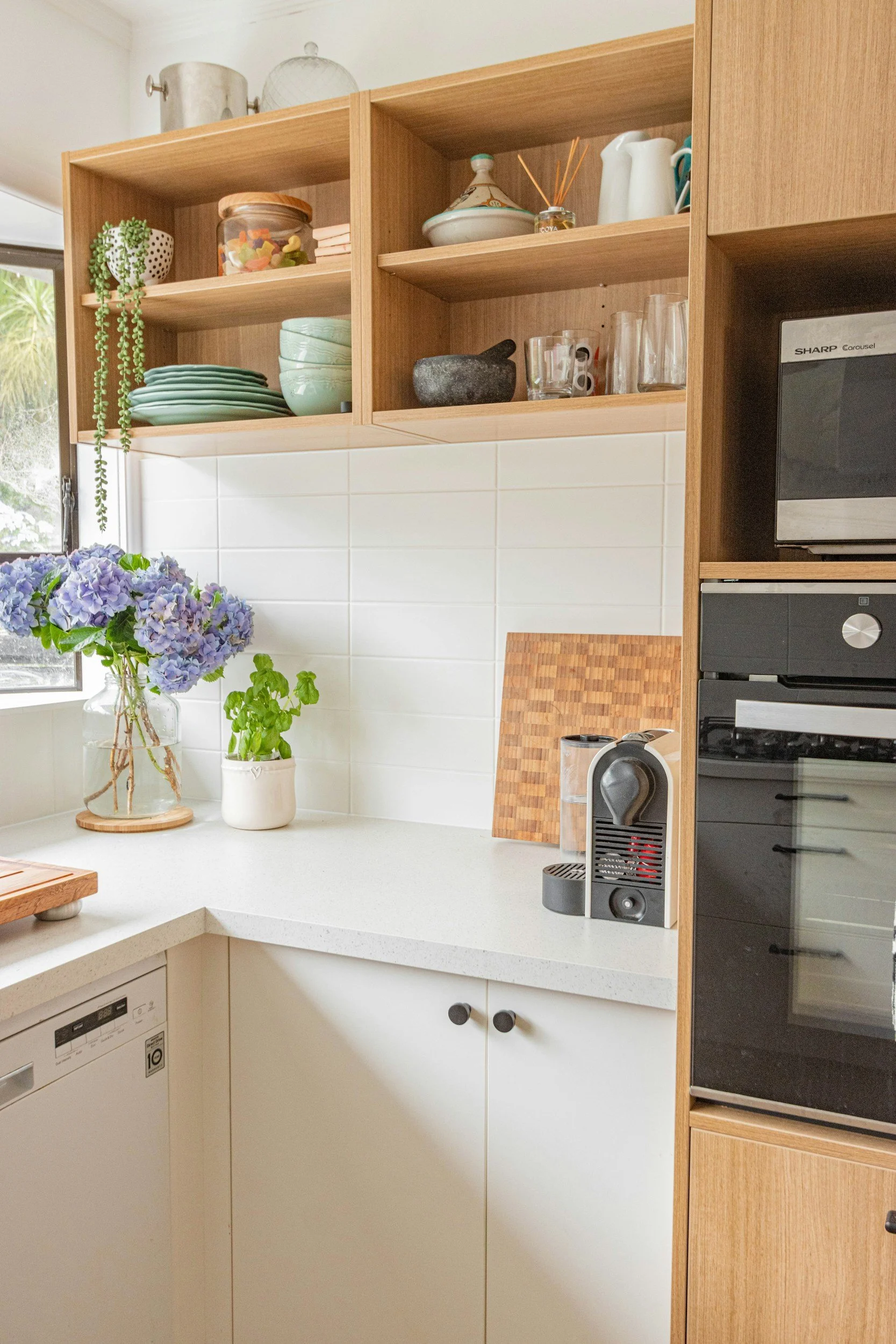 Kitchen corner with white countertops, open wooden shelves with dishes and glassware, a vase with purple flowers and green plants, a coffee machine, and built-in oven.