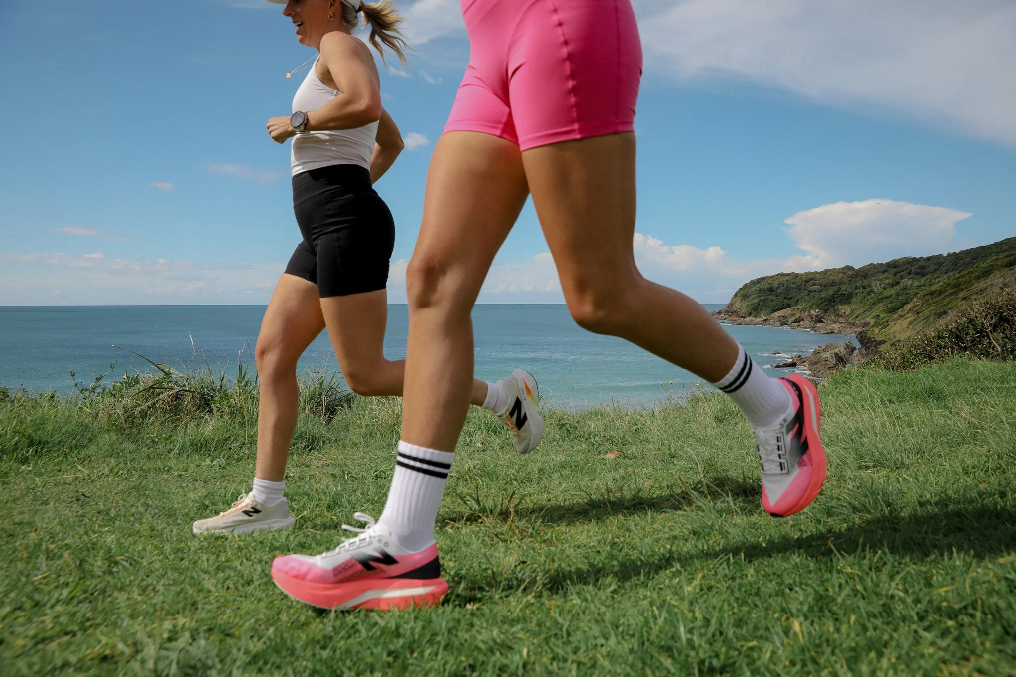 Two women running on grassy terrain near a coastline with ocean and sky in the background.