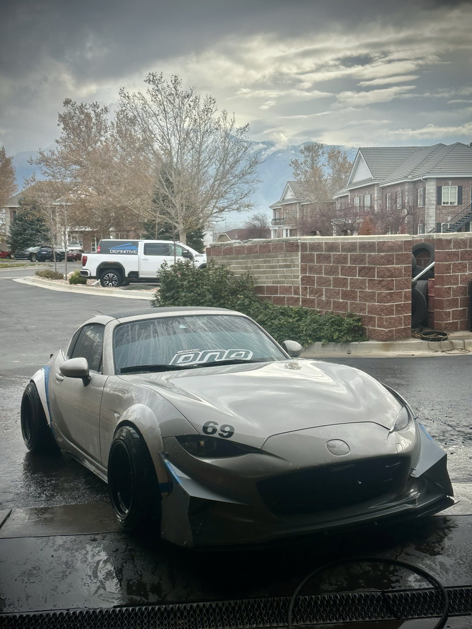 A silver sports car with black wheels parked on a wet surface, with a brick wall, trees, a white pickup truck, and residential buildings in the background on a cloudy day.