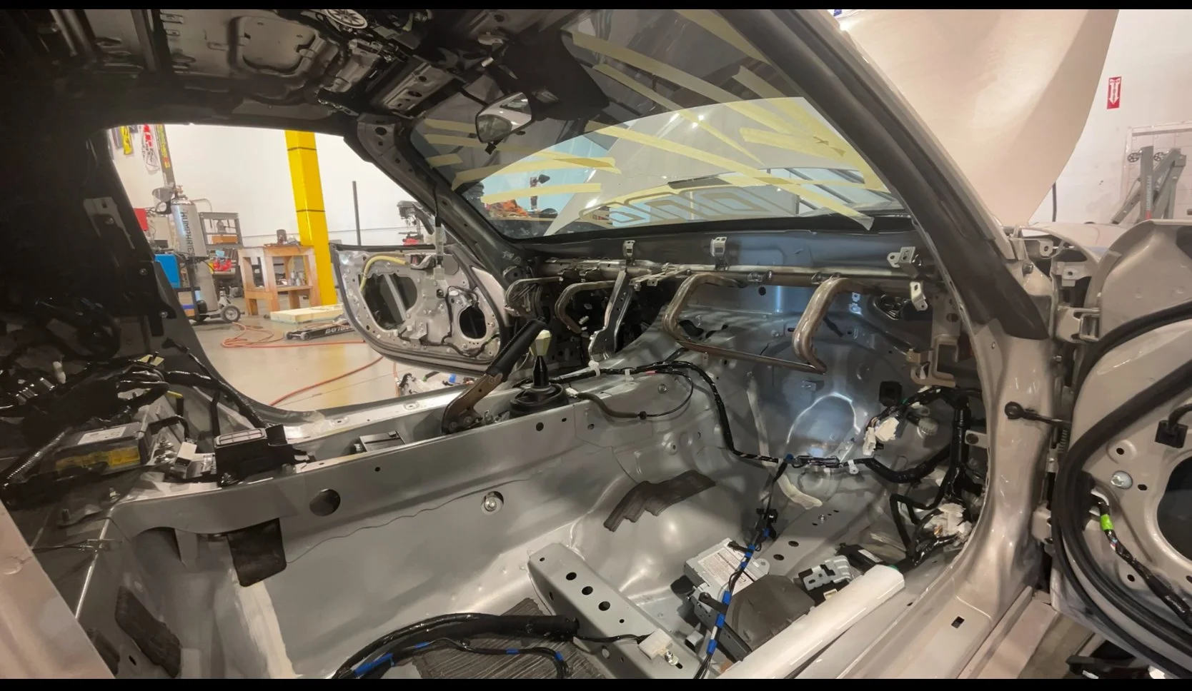 Interior view of a car with the dashboard and interior panels removed, showing the metal frame, wiring, and some mechanical components, in a workshop setting.