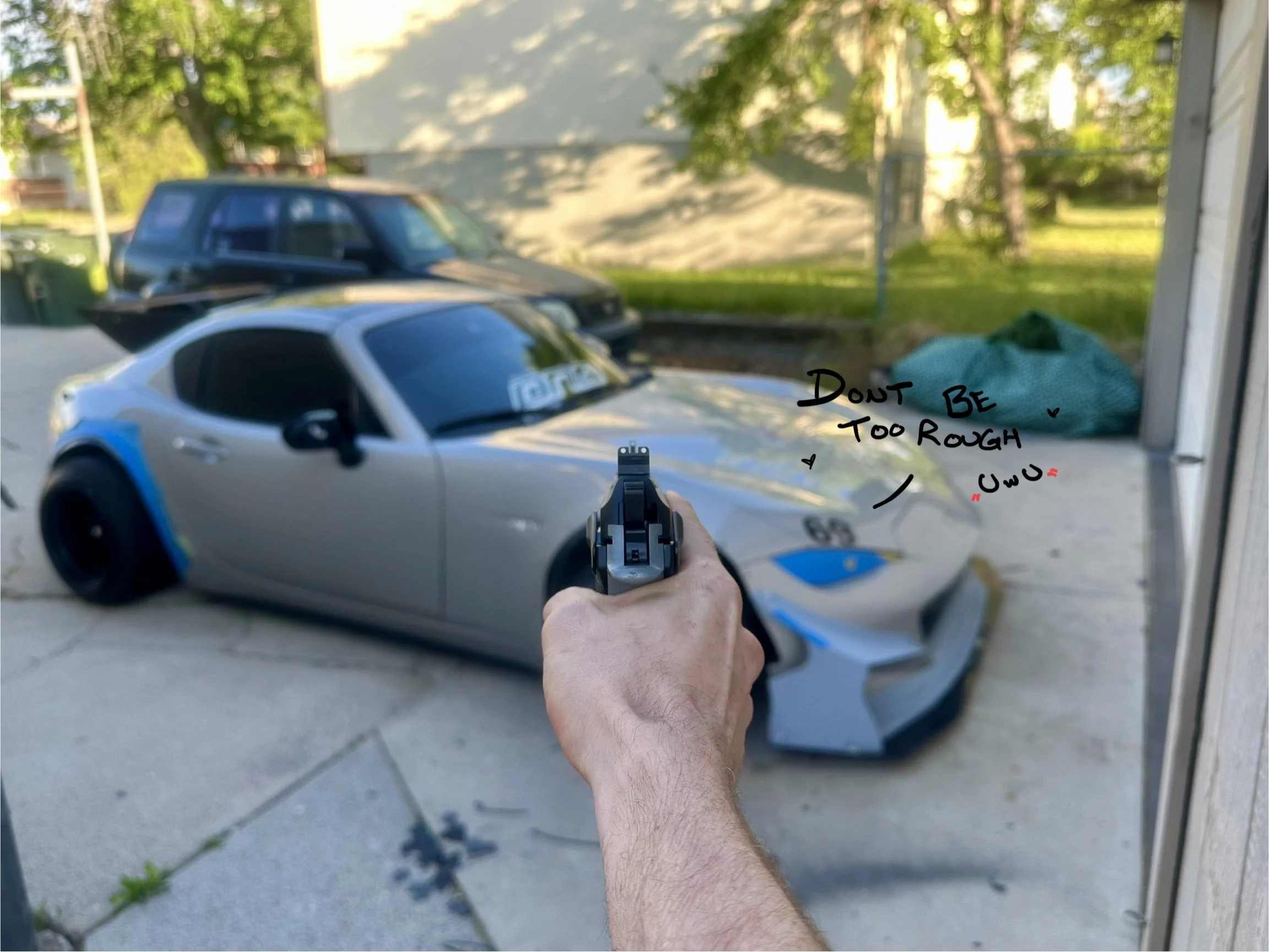 A person holding a gun in front of a scale model of a sports car with a handwritten message on the window that says 'Don't be too rough UwU'.