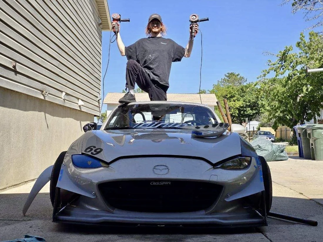 Person standing on top of a race car, holding power tools in each hand, with a house, trees, and a street in the background on a sunny day.