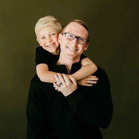 A smiling man with glasses is holding a young boy with blonde hair, both are wearing black and standing against a green background.