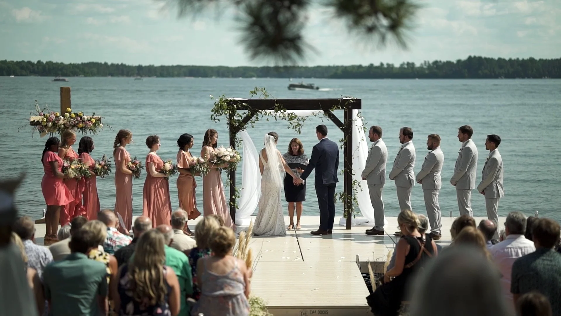 A wedding ceremony taking place outdoors by a lake, with the bride and groom exchanging vows under a wooden arch decorated with greenery and white fabric, surrounded by bridesmaids in pink dresses and groomsmen in white suits, with guests seated watching.