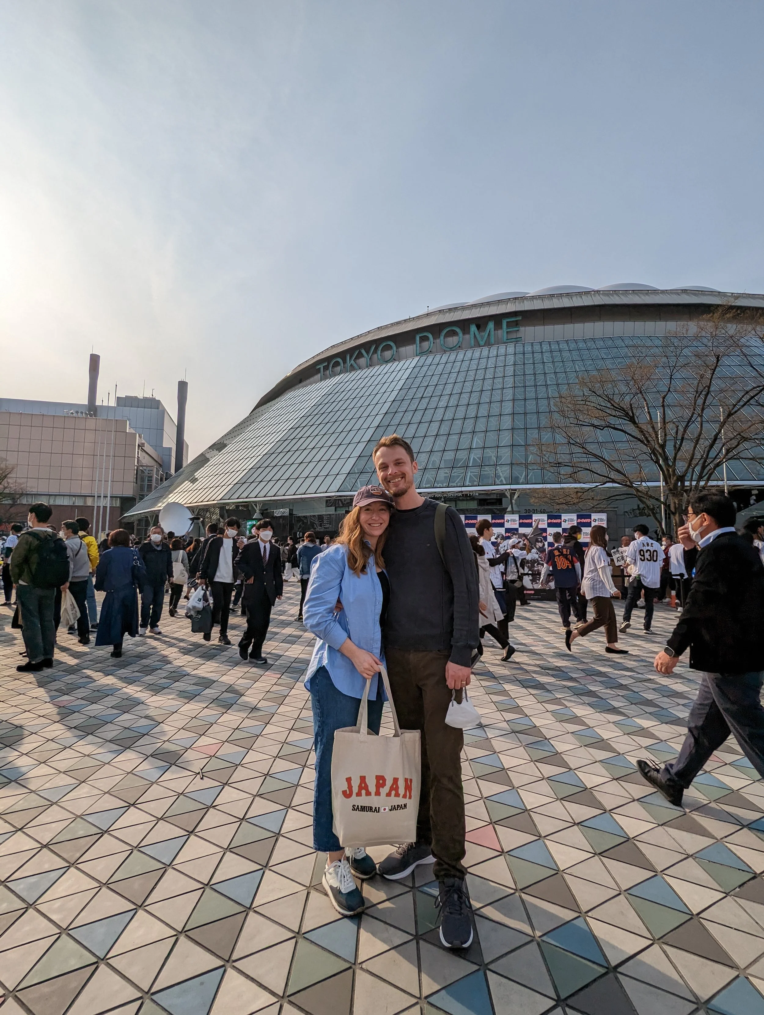 Two people standing in front of Tokyo Dome, smiling at the camera. The woman is holding a tote bag that says 'JAPAN' and is wearing a blue jacket and jeans. The man is wearing a black jacket and has his arm around the woman. There are many people walking around and some wearing masks in the background.
