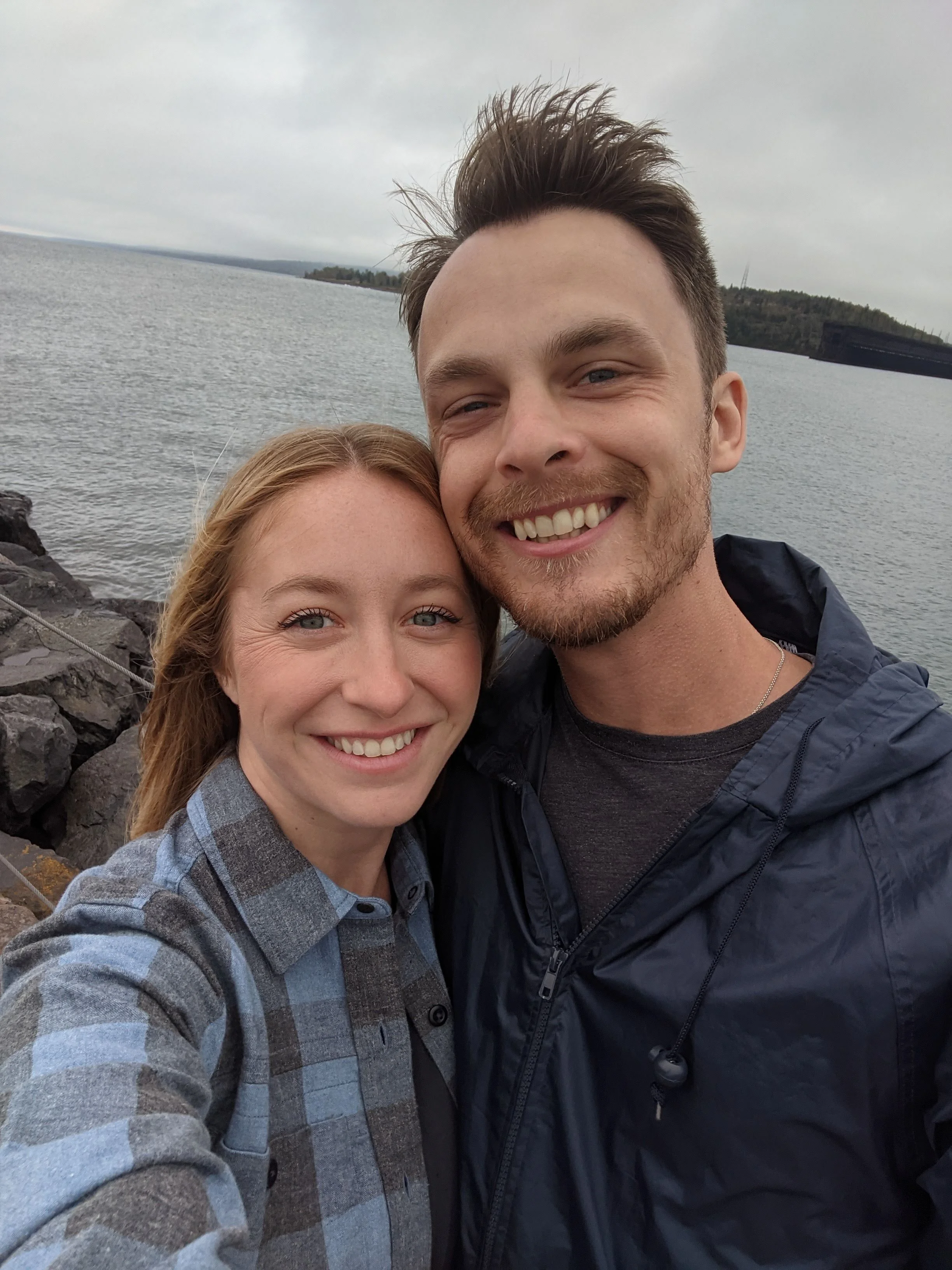 A smiling couple taking a selfie outdoors near a body of water with rocky shoreline and cloudy sky.