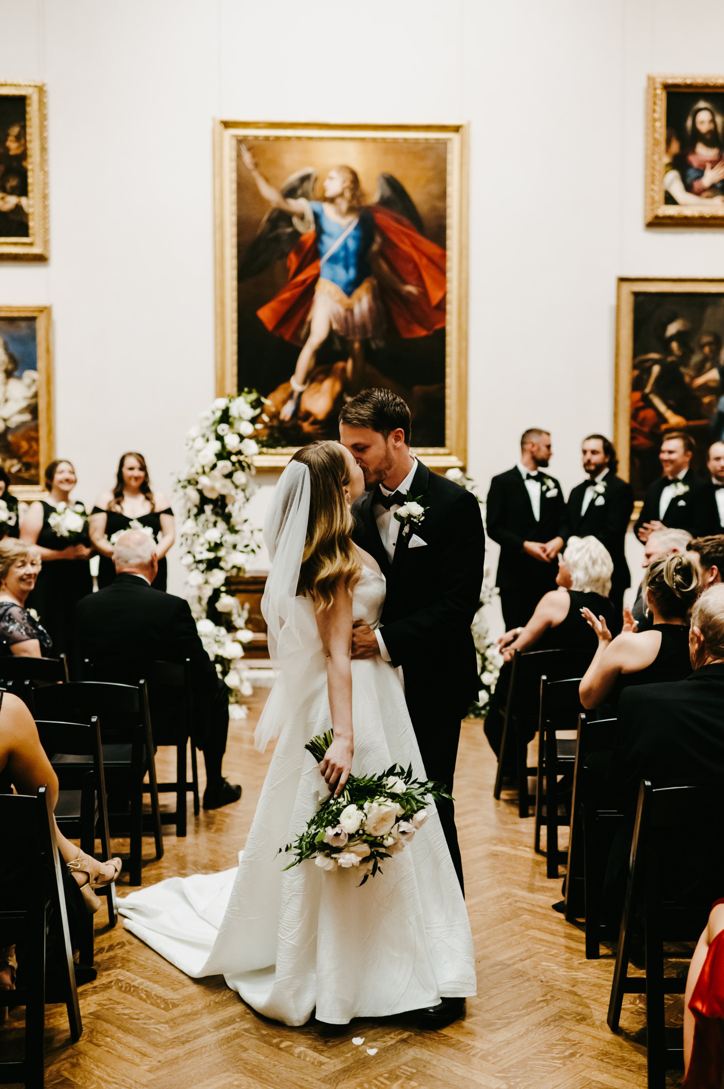 A newly married couple sharing a kiss during their wedding ceremony in an art gallery surrounded by guests and large classical paintings.
