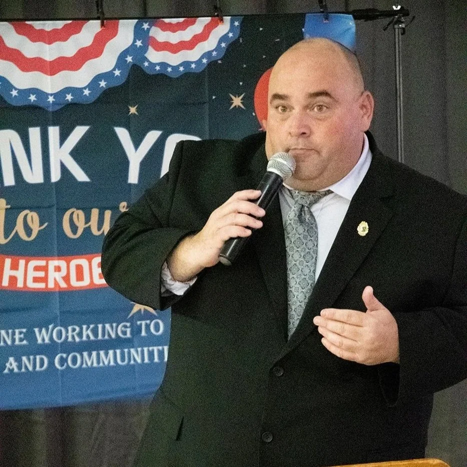 A man wearing a black suit, white shirt, and patterned tie speaking into a microphone at a community event with a patriotic themed backdrop.