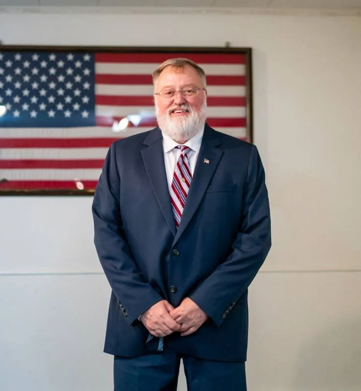 A man with glasses, a beard, and a suit standing indoors in front of an American flag.