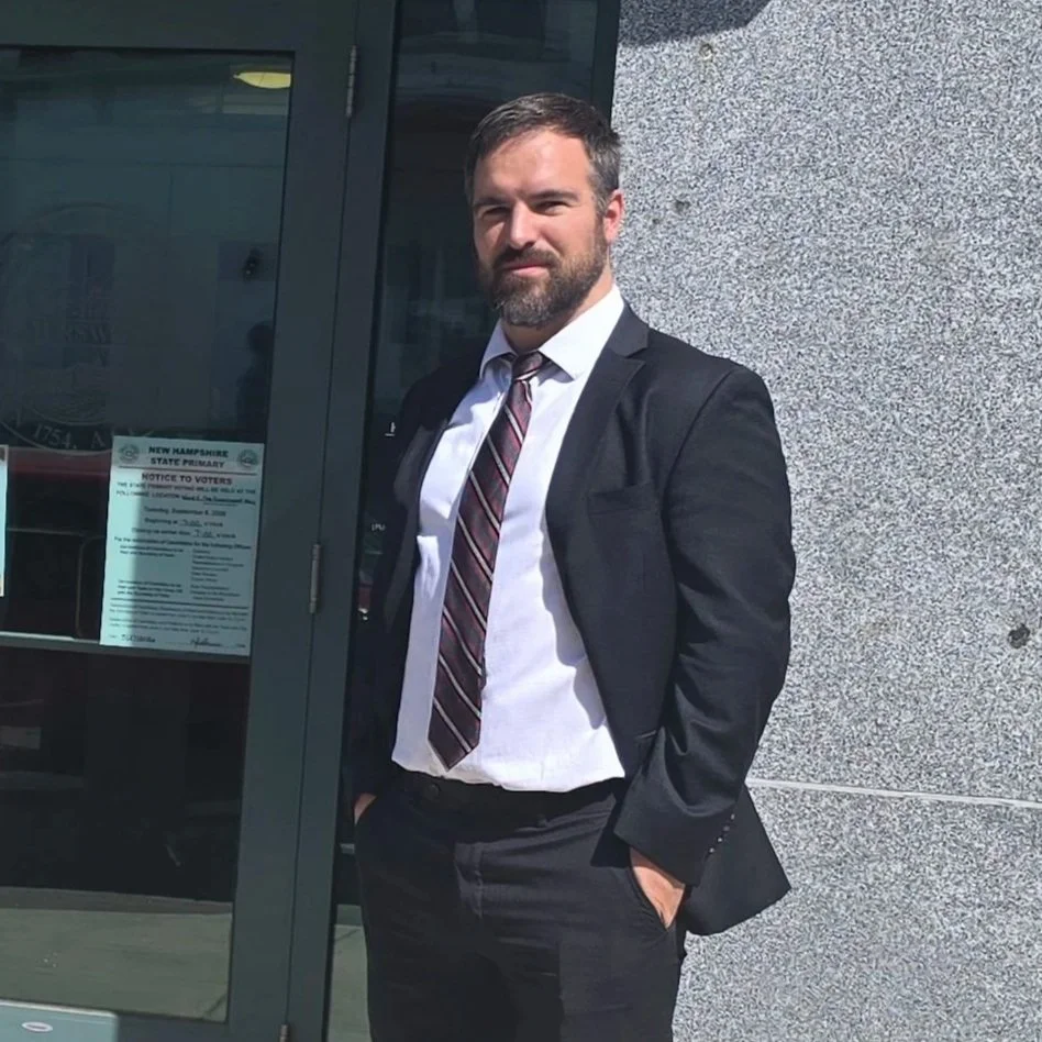 A man in a black suit, white shirt, and striped tie standing outside near a glass door with a gray stone wall behind him.