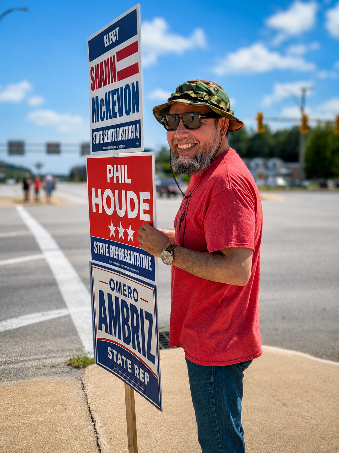 A man with a beard, wearing sunglasses, a camouflage hat, and a red T-shirt, standing outdoors and holding a political campaign sign supporting Shawn McKeown and Phil Houde with other candidates' names.