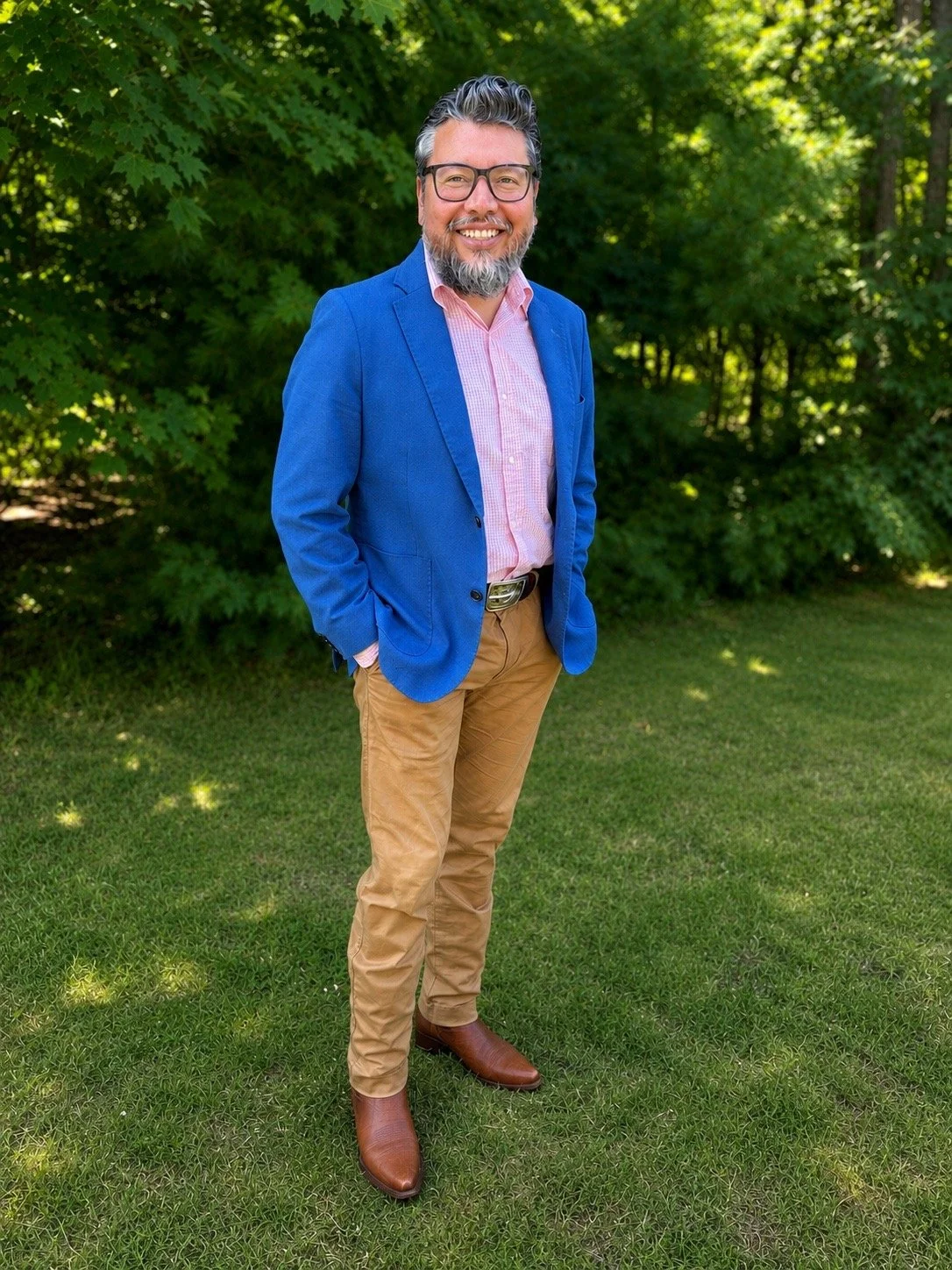 A man standing outdoors on grass with trees in the background, dressed in a bright blue blazer, pink button-up shirt, tan pants, and brown boots, smiling at the camera.