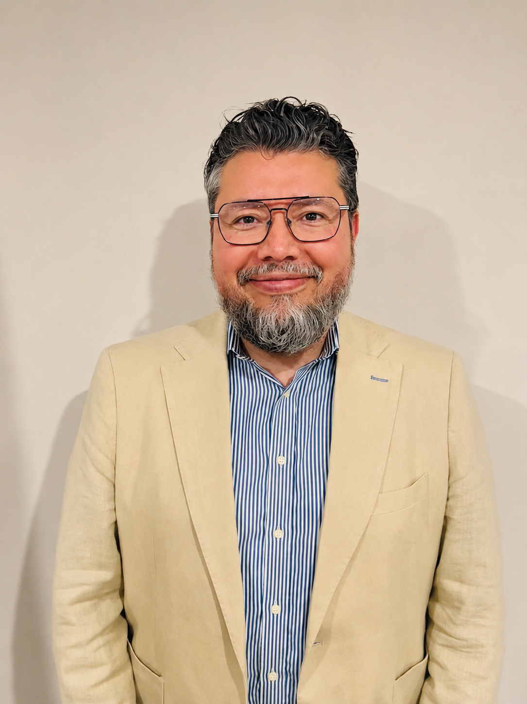 Man with glasses and a beard wearing a beige blazer and striped shirt, standing against a plain wall.