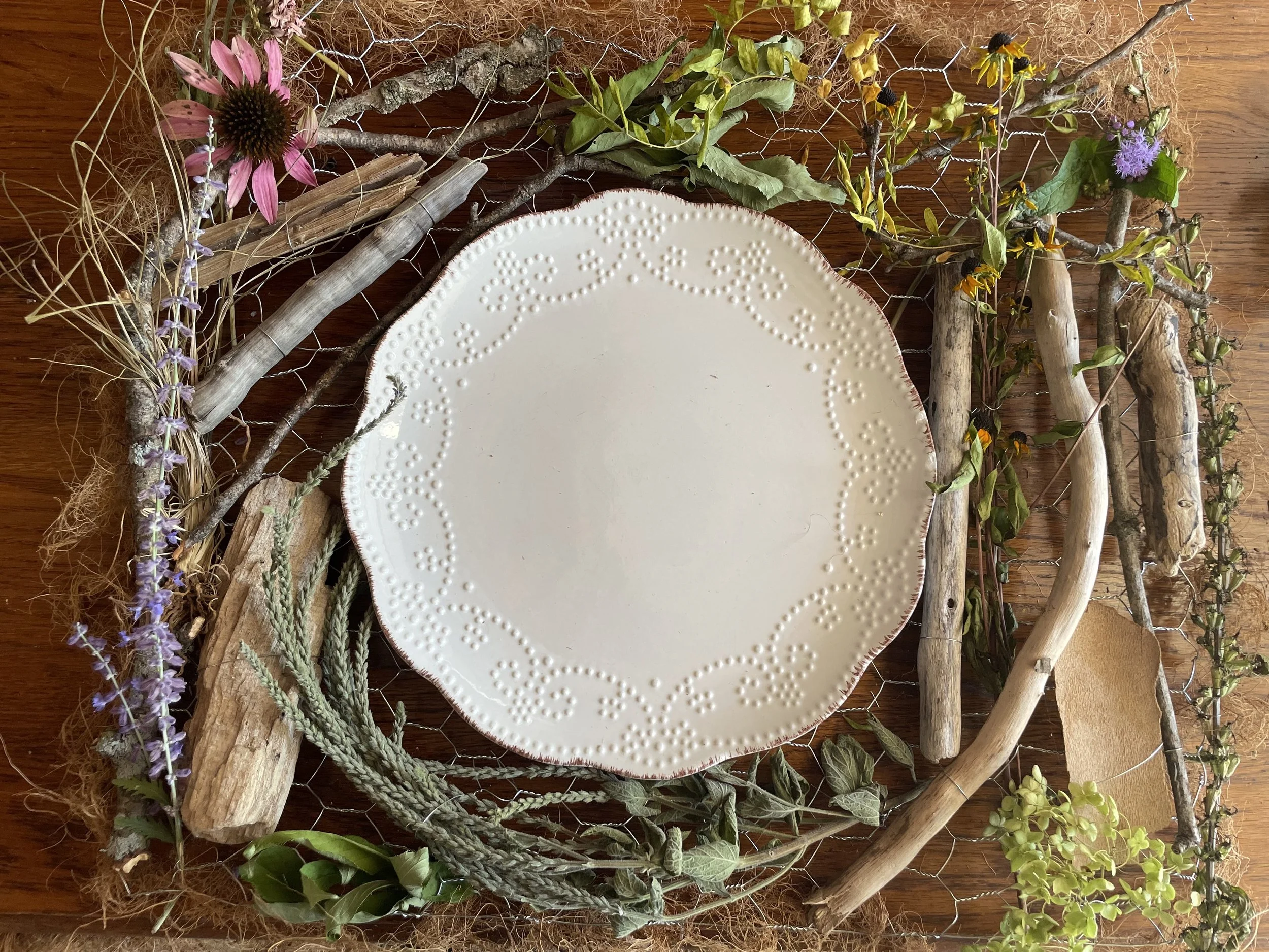 Decorative centerpiece with a white plate surrounded by a wreath of dried flowers, branches, driftwood, and greenery on a wooden surface.