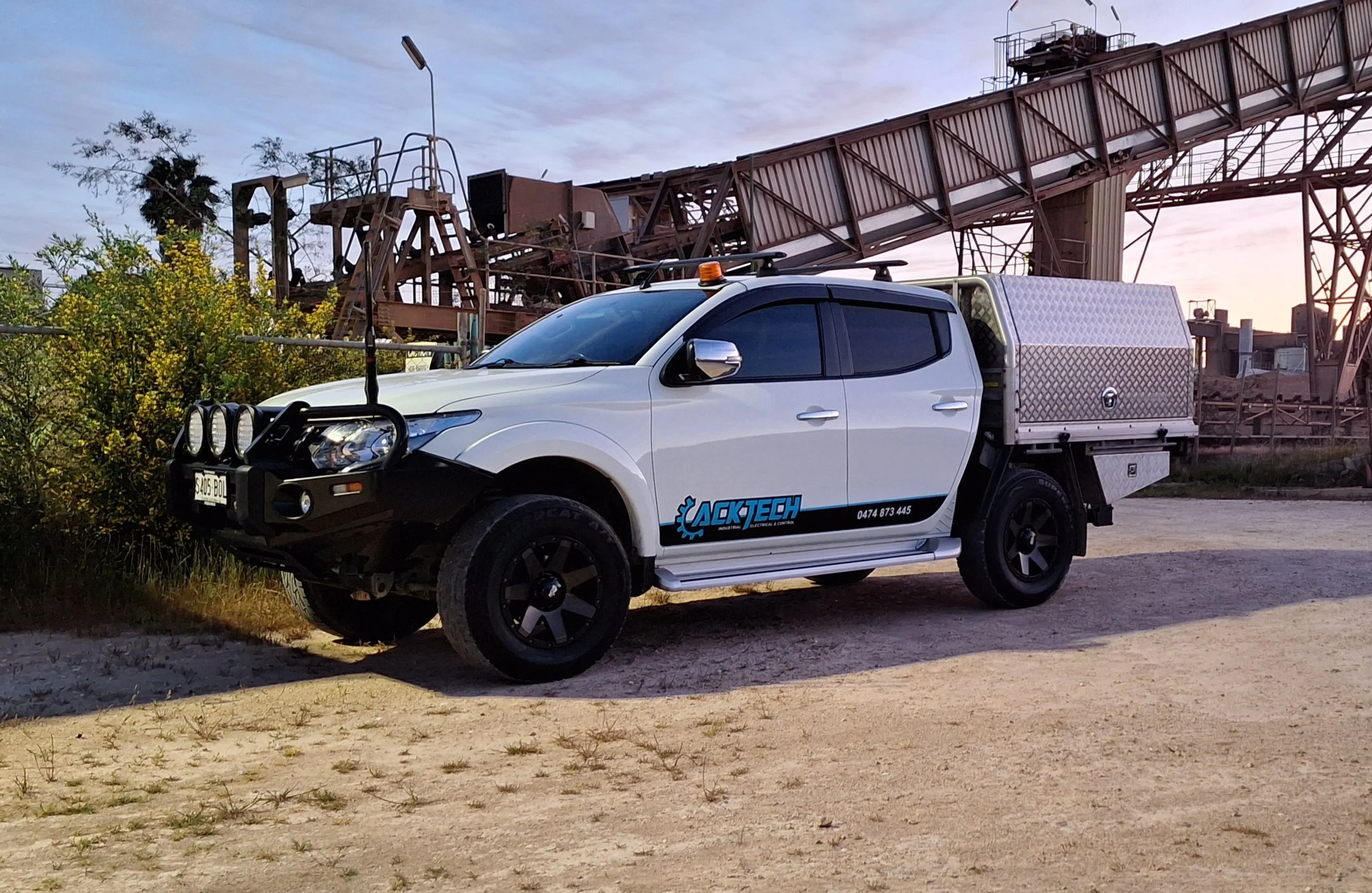 A white utility vehicle with a company logo and contact information on the side, parked on a dirt surface beside a bush. The vehicle has black wheels, roof racks with a flashing light, and a metallic storage box in the bed. In the background, there is an industrial structure with rusted metal beams and conveyor belts, under a partly cloudy sky.
