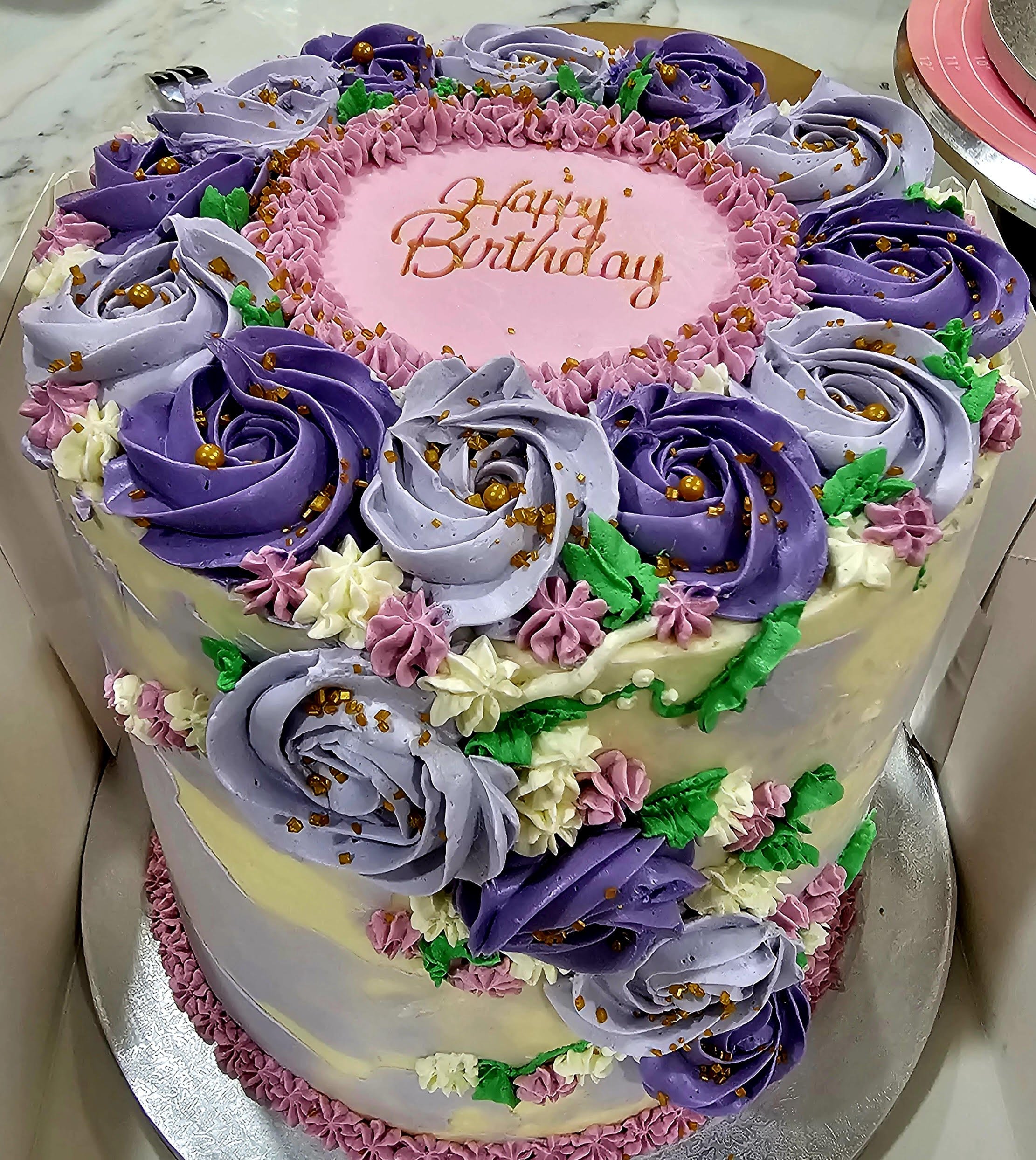 A two-tiered birthday cake decorated with purple, pink, white, and green floral icing, with a pink circular message in the center that reads 'Happy Birthday' outlined in a darker pink.