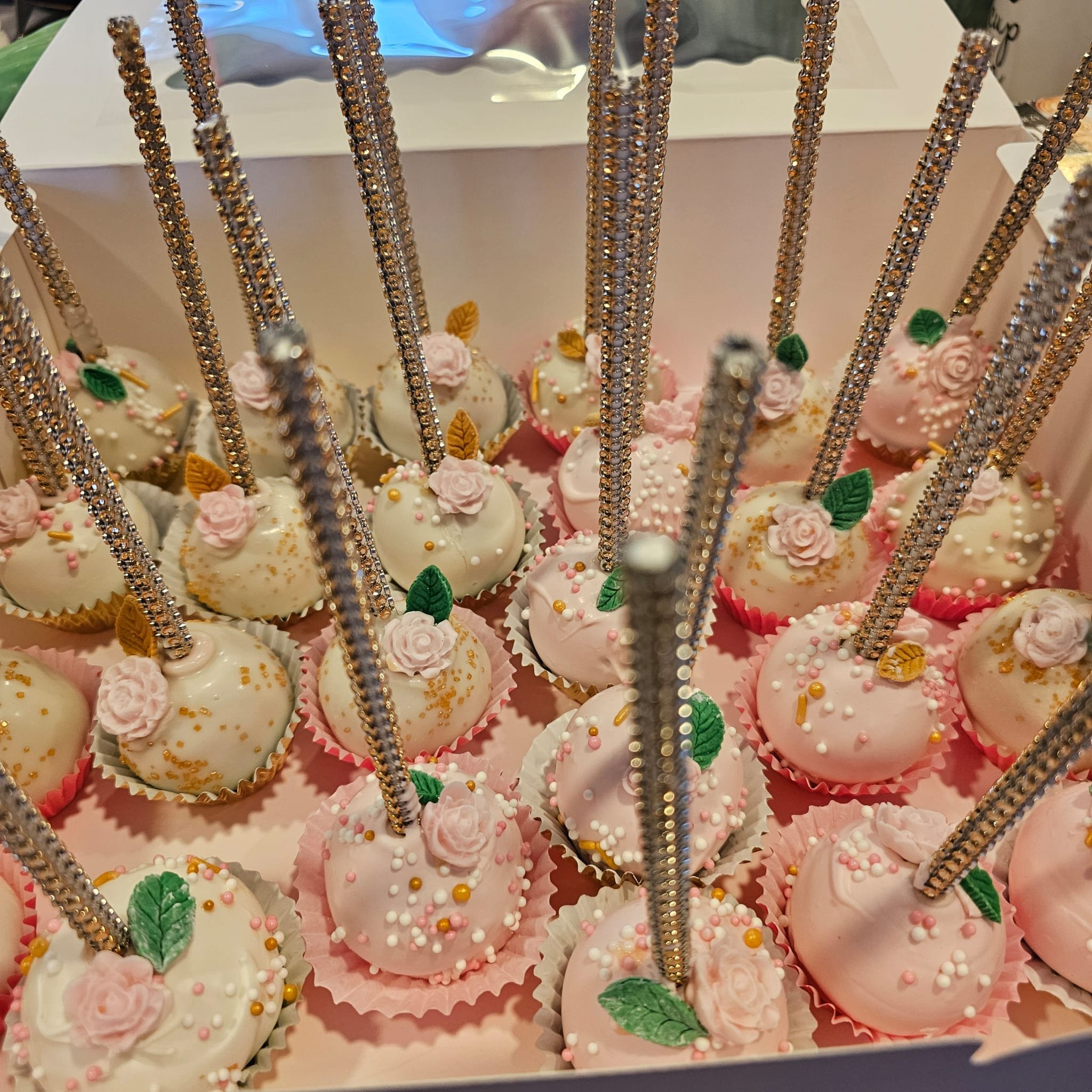 Decorated cake pops with pink and white icing, adorned with small pink roses, green leaves, and colorful sprinkles, inside a box with tall, glittery, gold sticks.