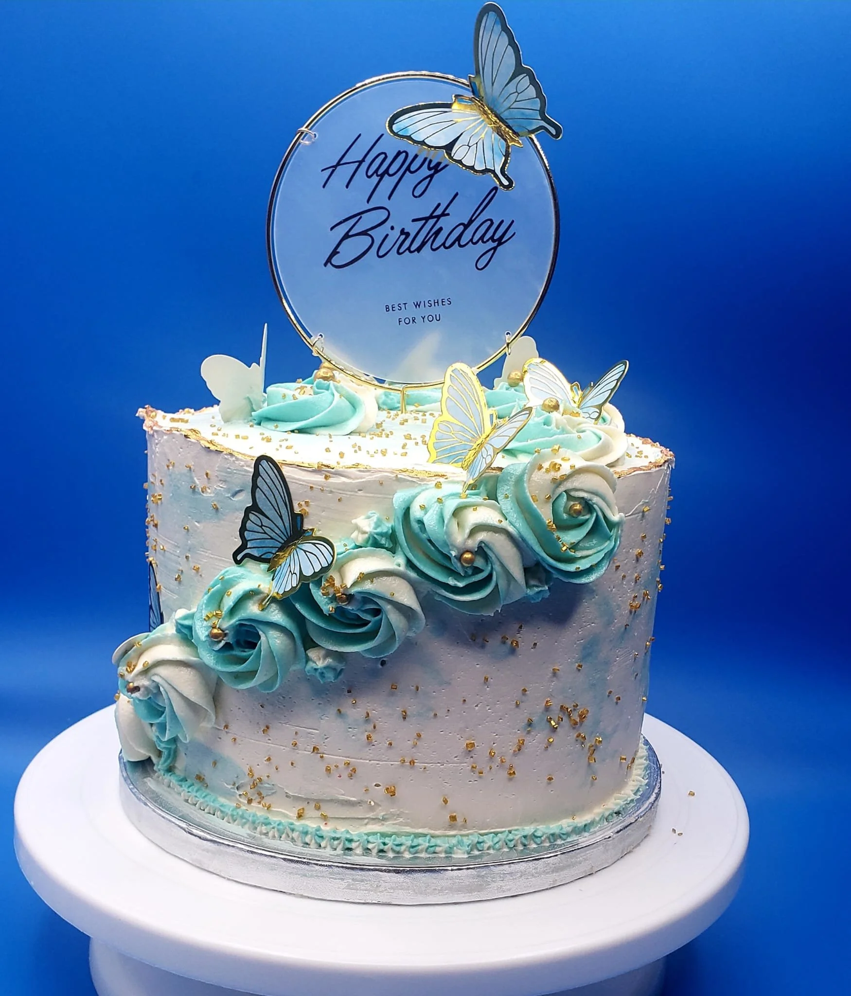 A birthday cake decorated with light blue and white swirls and butterflies, gold accents, and a circular topper that says 'Happy Birthday' with butterflies, the top is light blue with butterflies, and the background is a solid blue color.